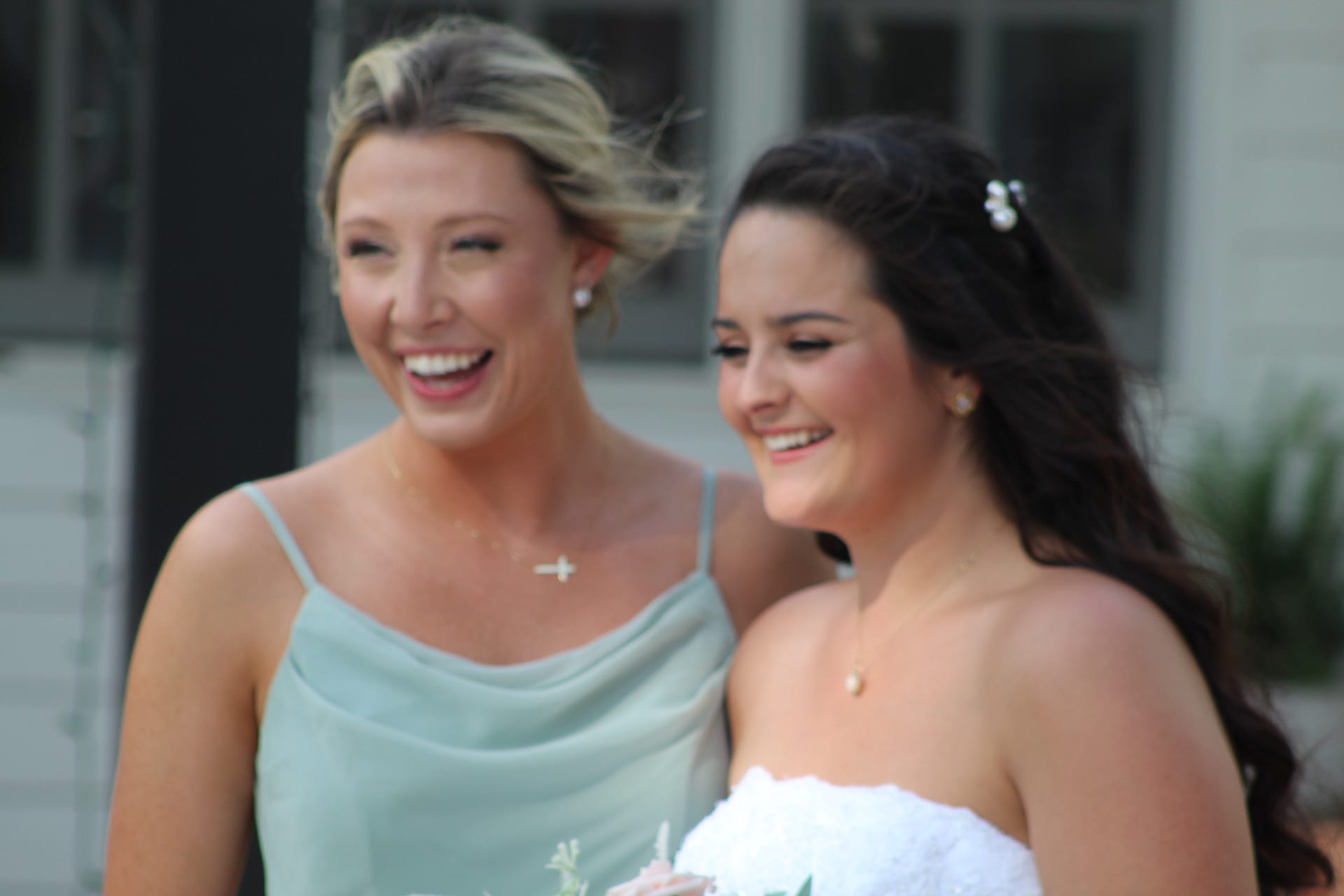 A laughing bride in white and her bridesmaid in sage green share a joyful candid moment outdoors at Scarborough House.