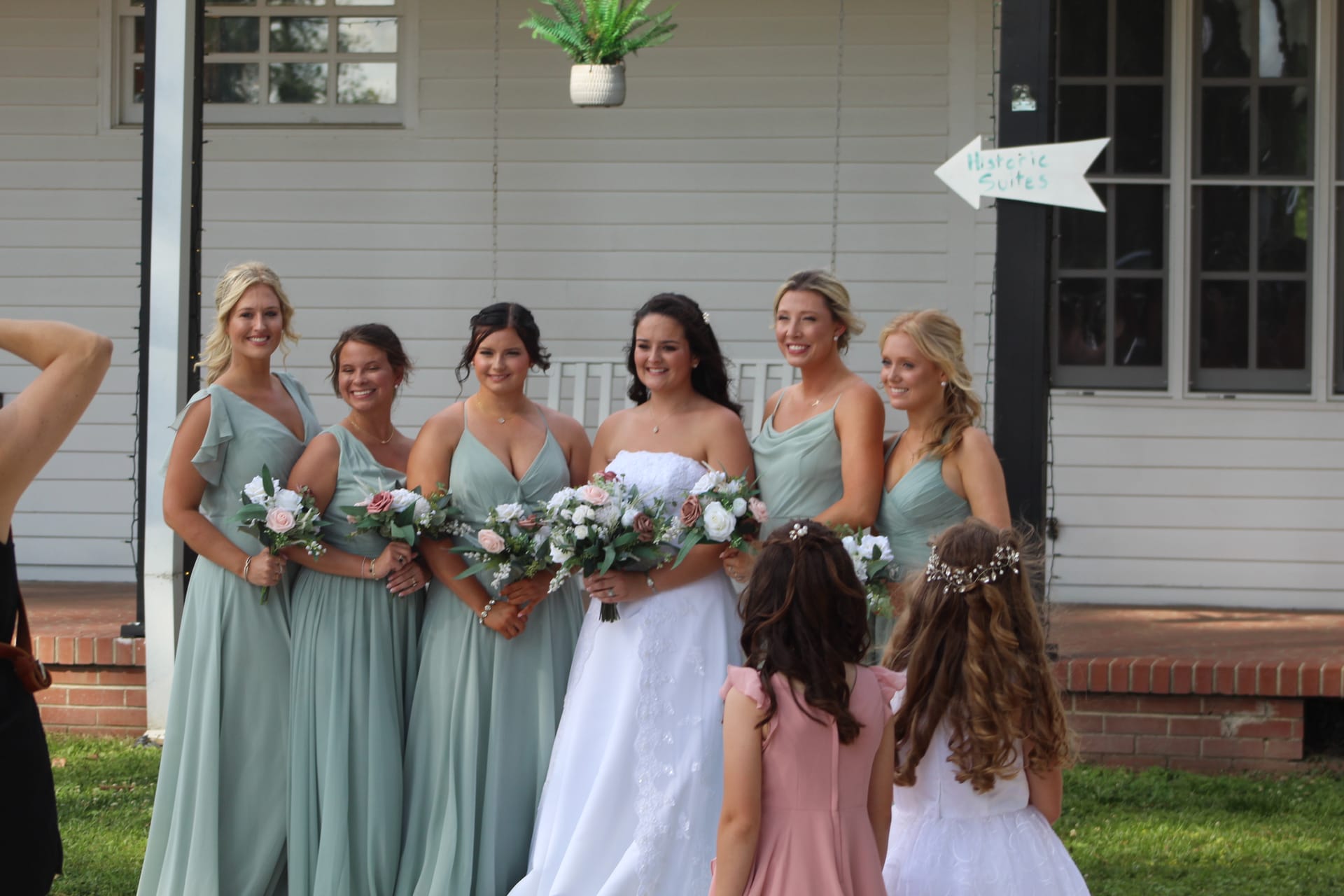 Bride in white gown surrounded by five bridesmaids in sage green dresses holding floral bouquets outside a historic white manor house with two flower girls in the foreground.