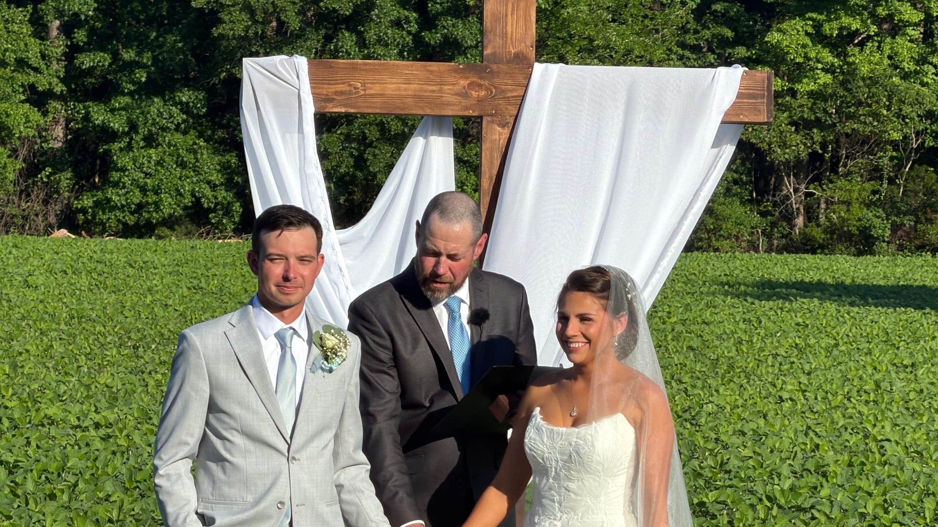A bride in a white lace gown and groom in a light gray suit exchange vows before a rustic wooden cross draped in white fabric with green farmland fields in the background at an outdoor wedding ceremony.