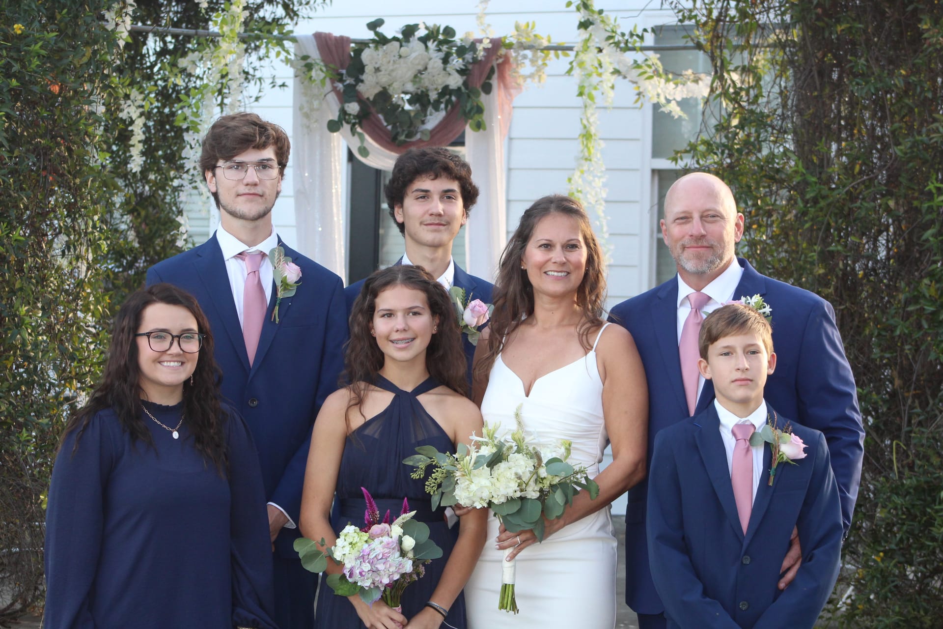 Bride in white gown and groom in navy suit pose with five children in coordinated navy and pink attire beneath a floral arch at Scarborough House historic estate.