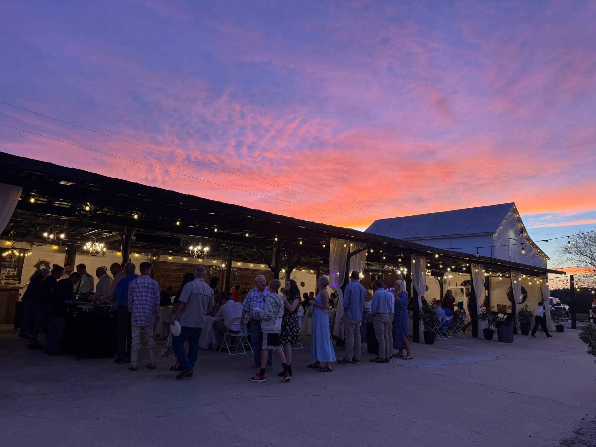 Guests mingle under string lights and chandeliers at an outdoor covered pavilion reception at Scarborough House as a vivid pink and purple sunset fills the evening sky.