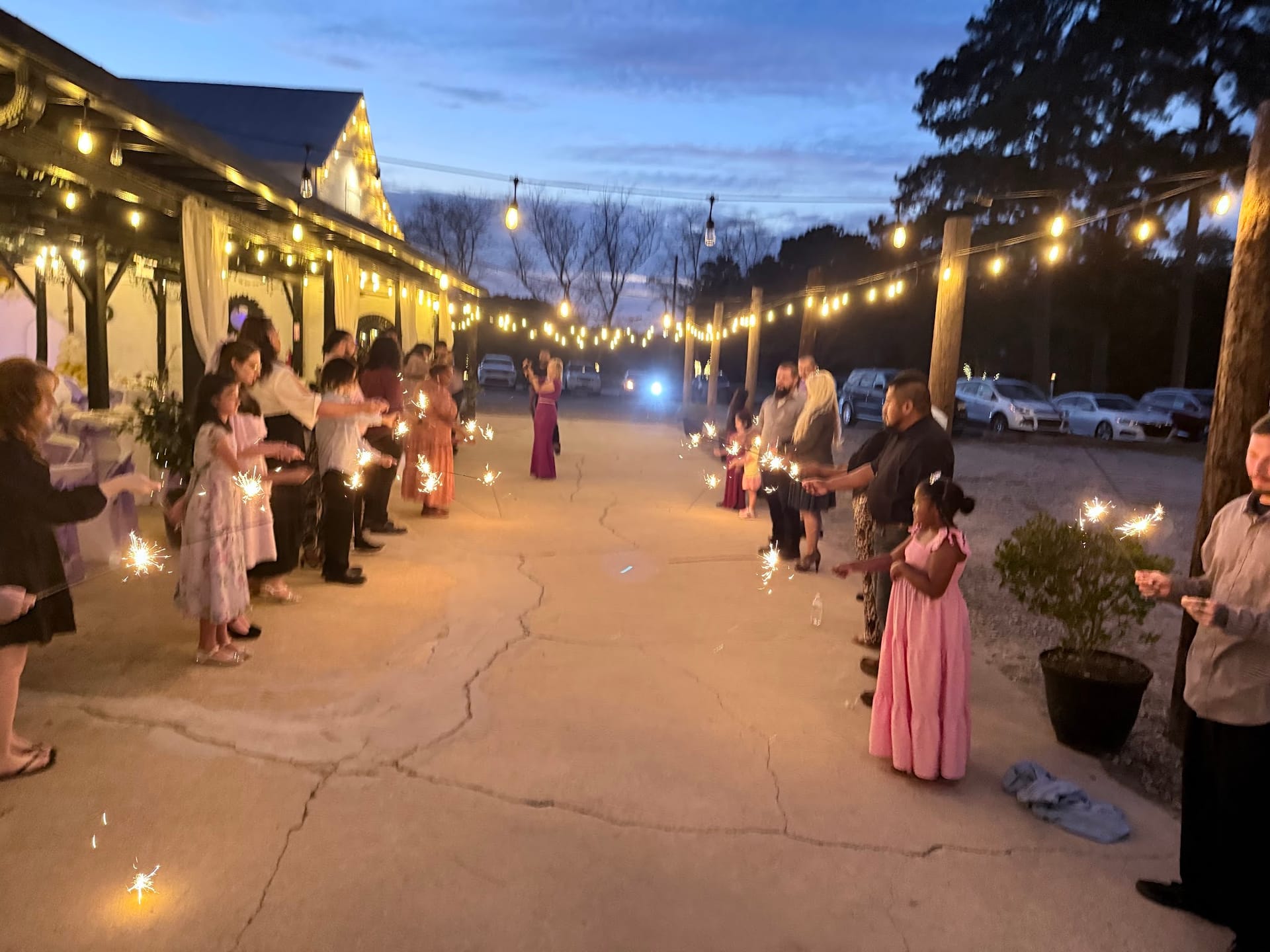 Wedding guests young and old line a glowing pathway holding sparklers at dusk beneath string lights at Scarborough House as the couple walks through.