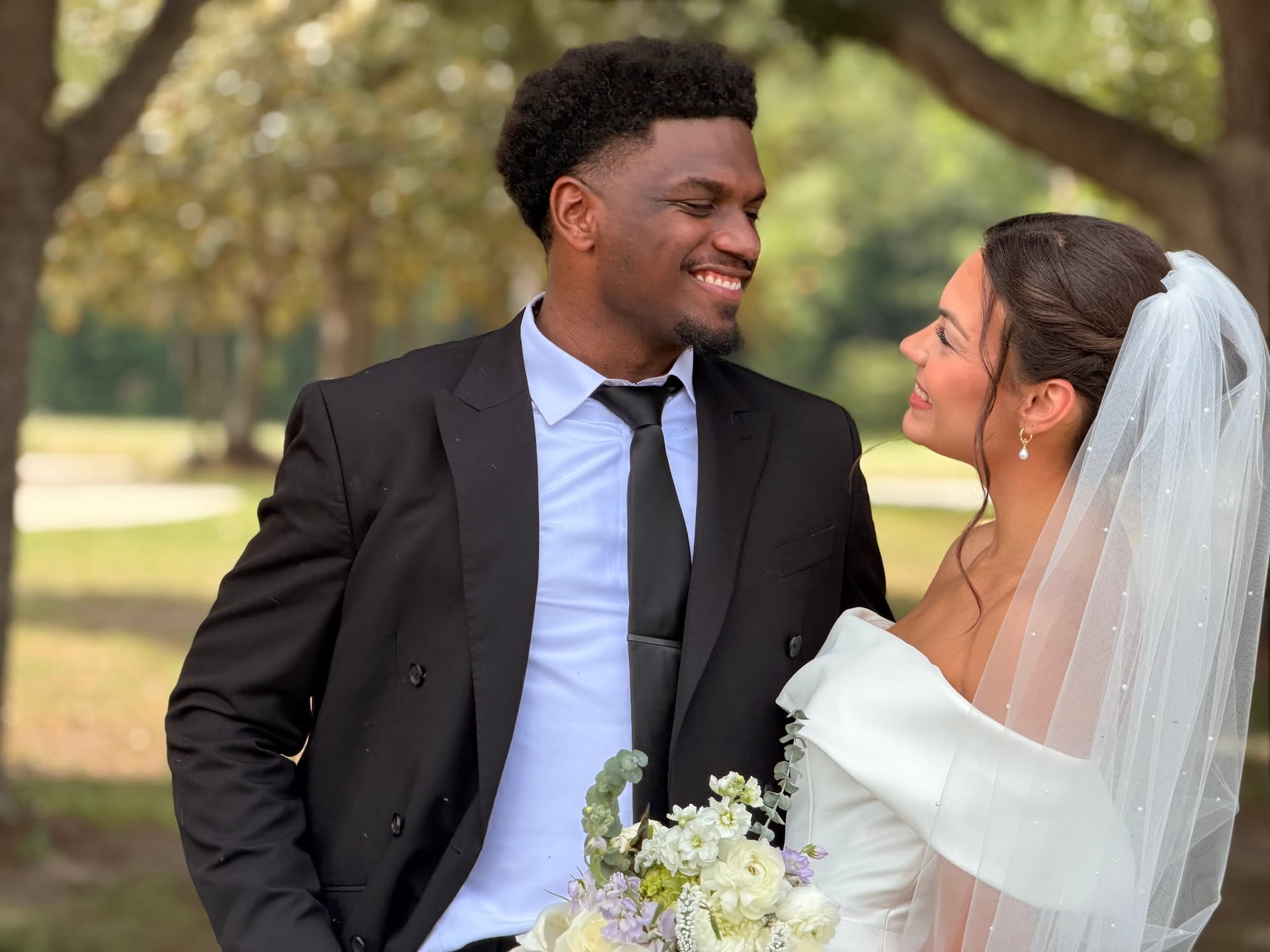 Bride in a white off-shoulder gown with veil holding a white floral bouquet gazes up at her smiling groom in a black suit beneath the oak trees at Scarborough House estate.