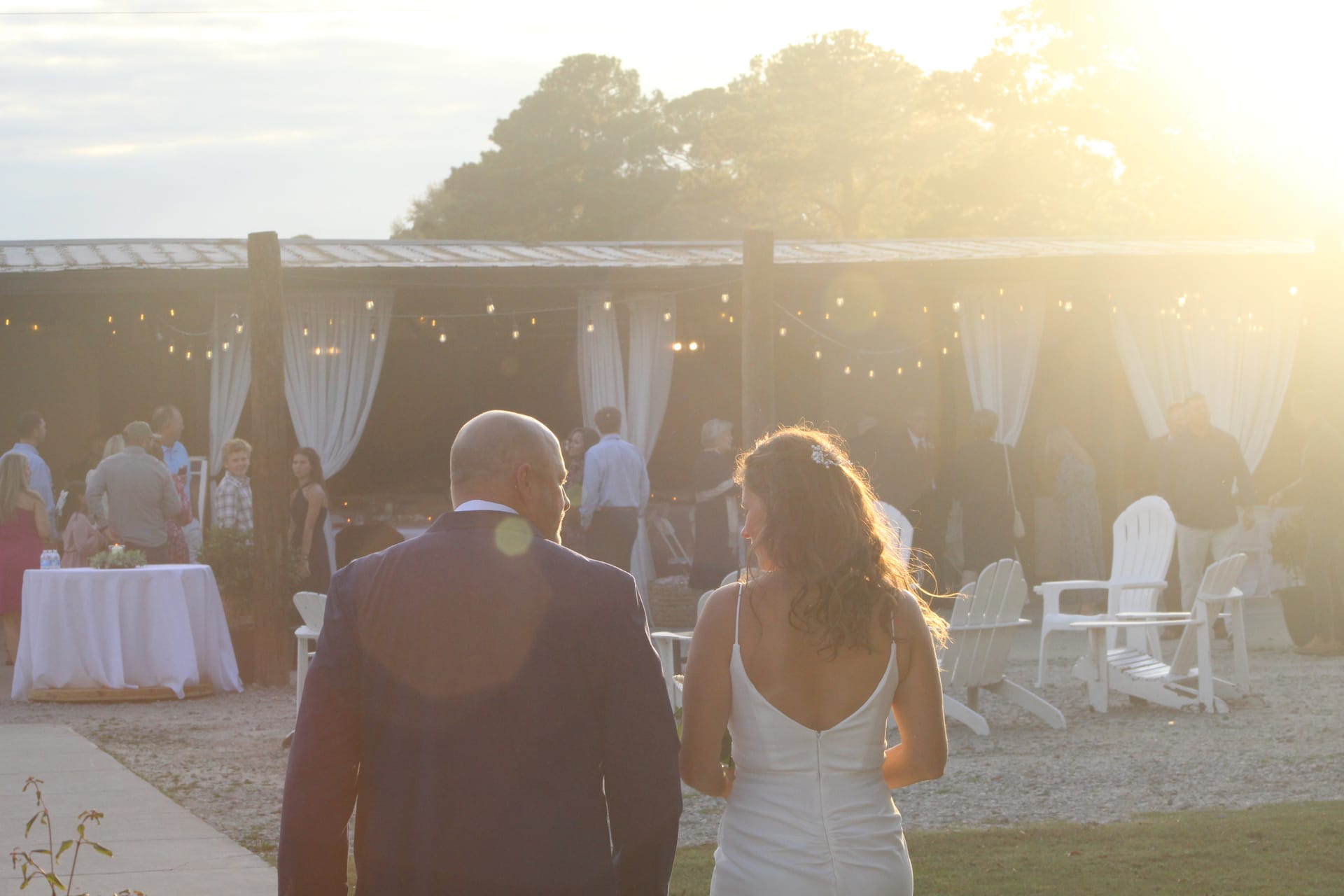 Bride and groom walk hand in hand toward a string-lit pavilion draped in white curtains at Scarborough House as a warm golden sunset bathes the estate in glowing light.