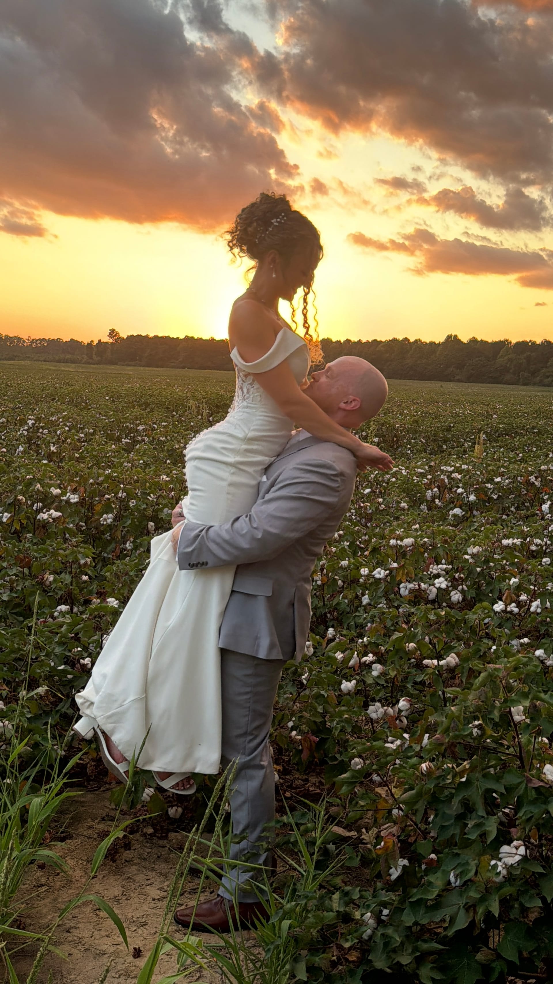 Groom in a grey suit lifts his bride in a white off-shoulder gown as they share a kiss amid a blooming cotton field beneath a vivid orange and gold sunset sky.