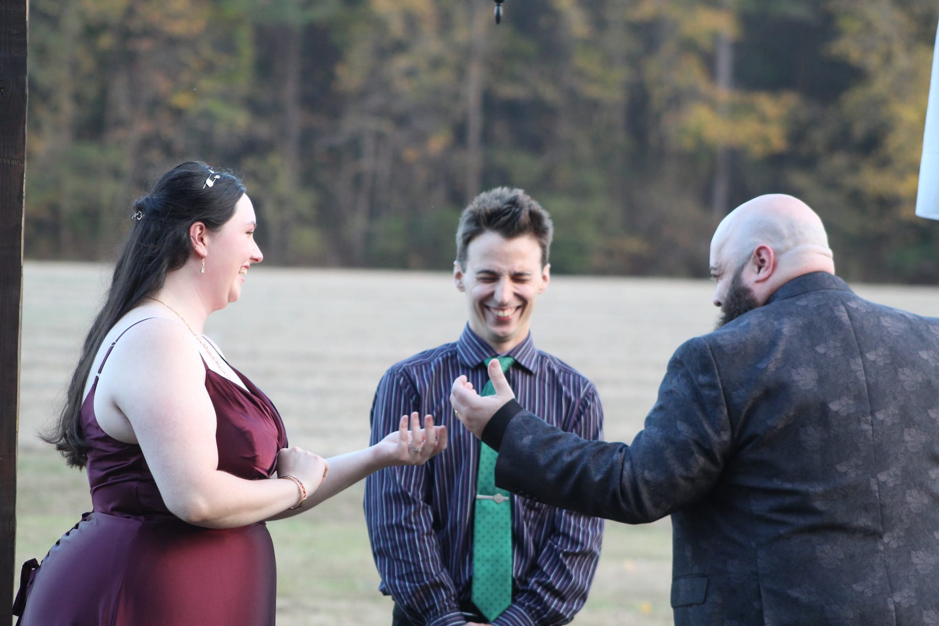 A smiling bride in a burgundy gown extends her hand to receive a ring from the groom in a dark floral suit while the officiant looks on happily during an outdoor ceremony at Scarborough House.