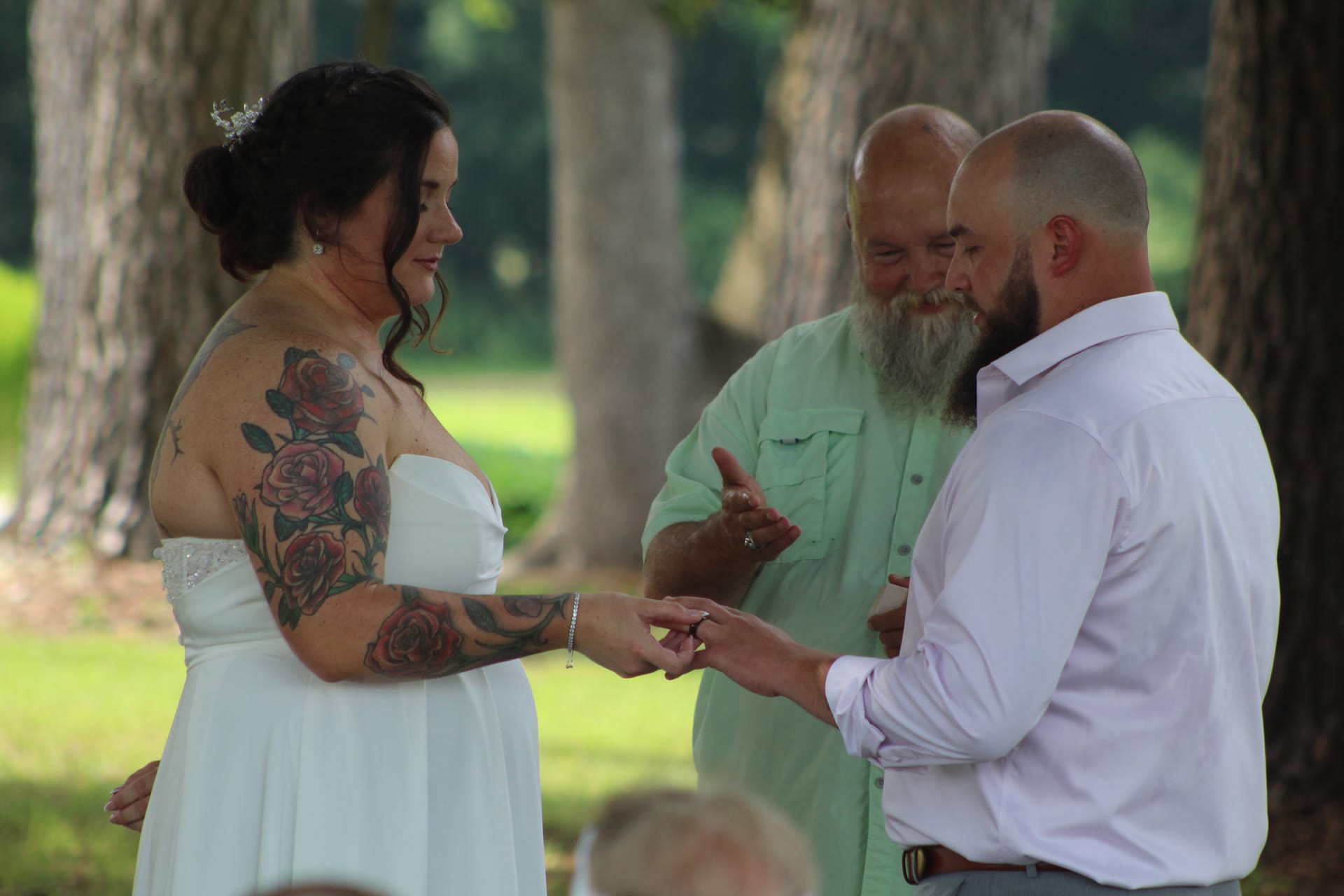 Groom places a wedding ring on his bride's finger during an outdoor ceremony beneath oak trees at Scarborough House while a bearded officiant looks on warmly.