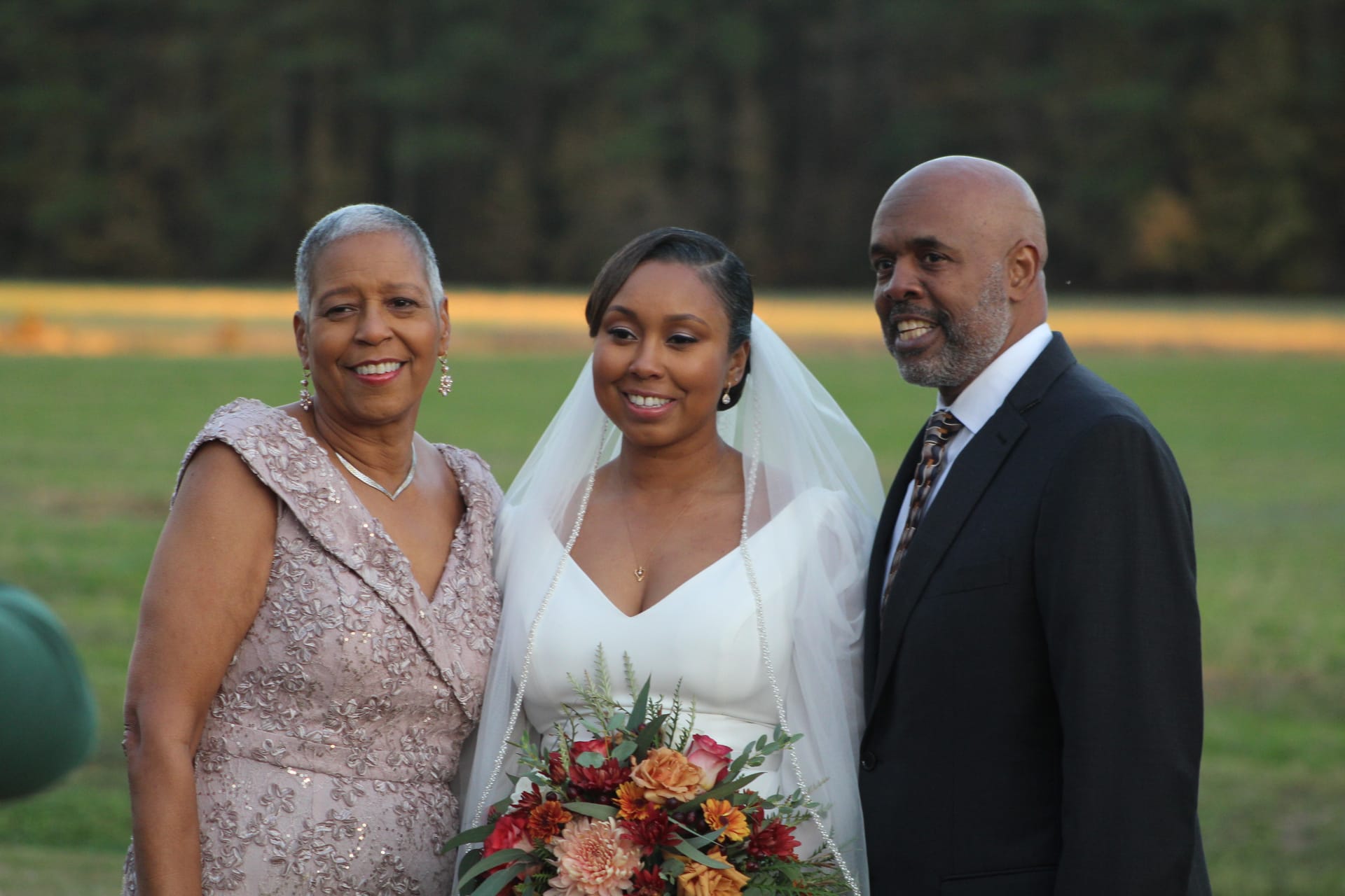 Bride in white gown and veil holding an autumn bouquet poses and smiles with her mother in a blush gown and her suited father on the ceremony lawn at Scarborough House.
