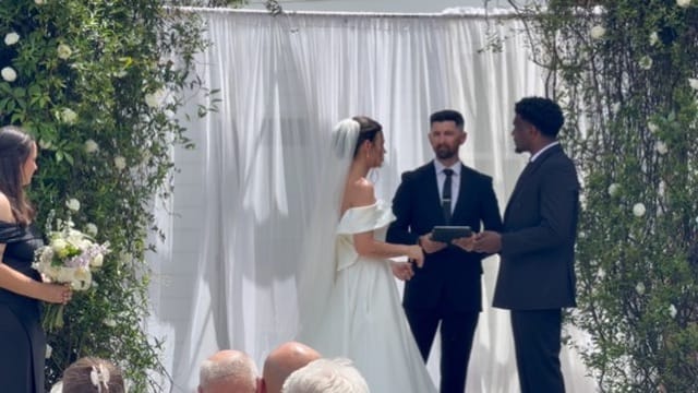 Bride and groom exchange vows beneath a lush floral and greenery arch with white draped backdrop at an outdoor wedding ceremony.