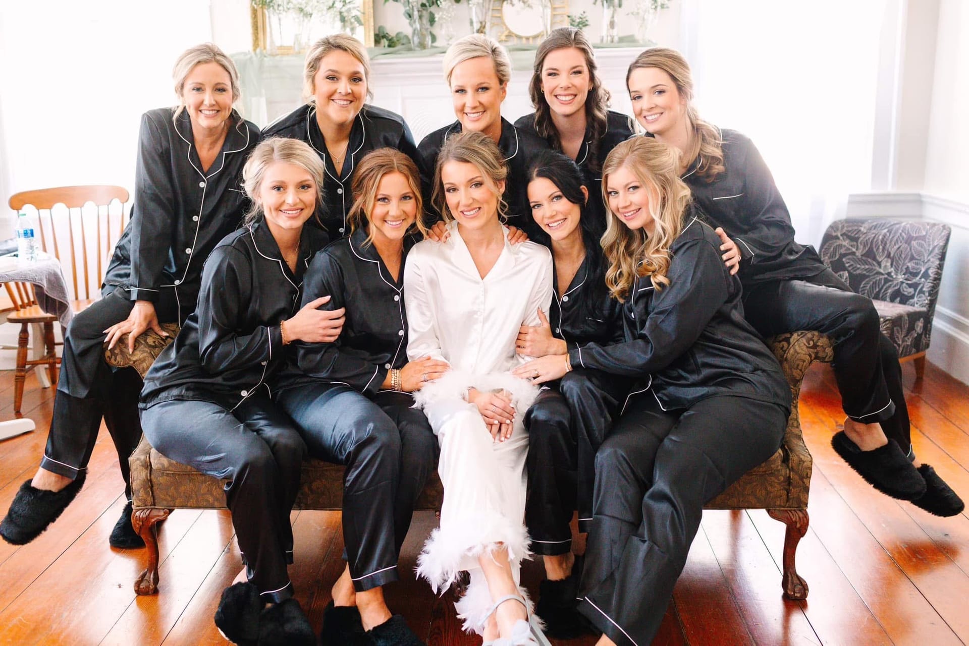 Bride in white satin robe surrounded by nine bridesmaids wearing matching black satin pajamas seated together on a vintage settee during wedding morning preparations at Scarborough House.
