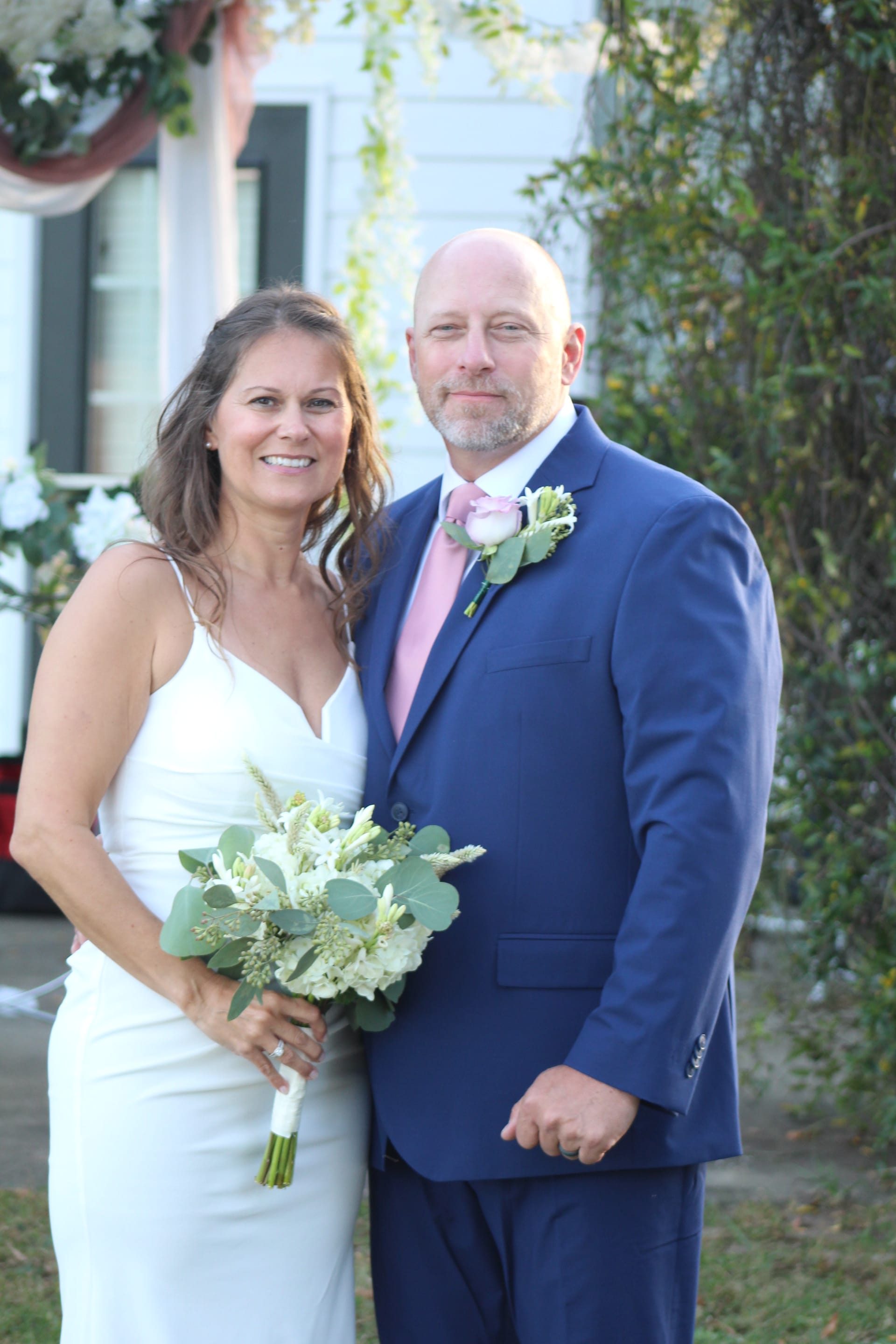 Bride in a white gown holding a white and greenery bouquet stands beside her groom in a navy suit with a pink tie and rose boutonniere in front of the historic Scarborough House estate.