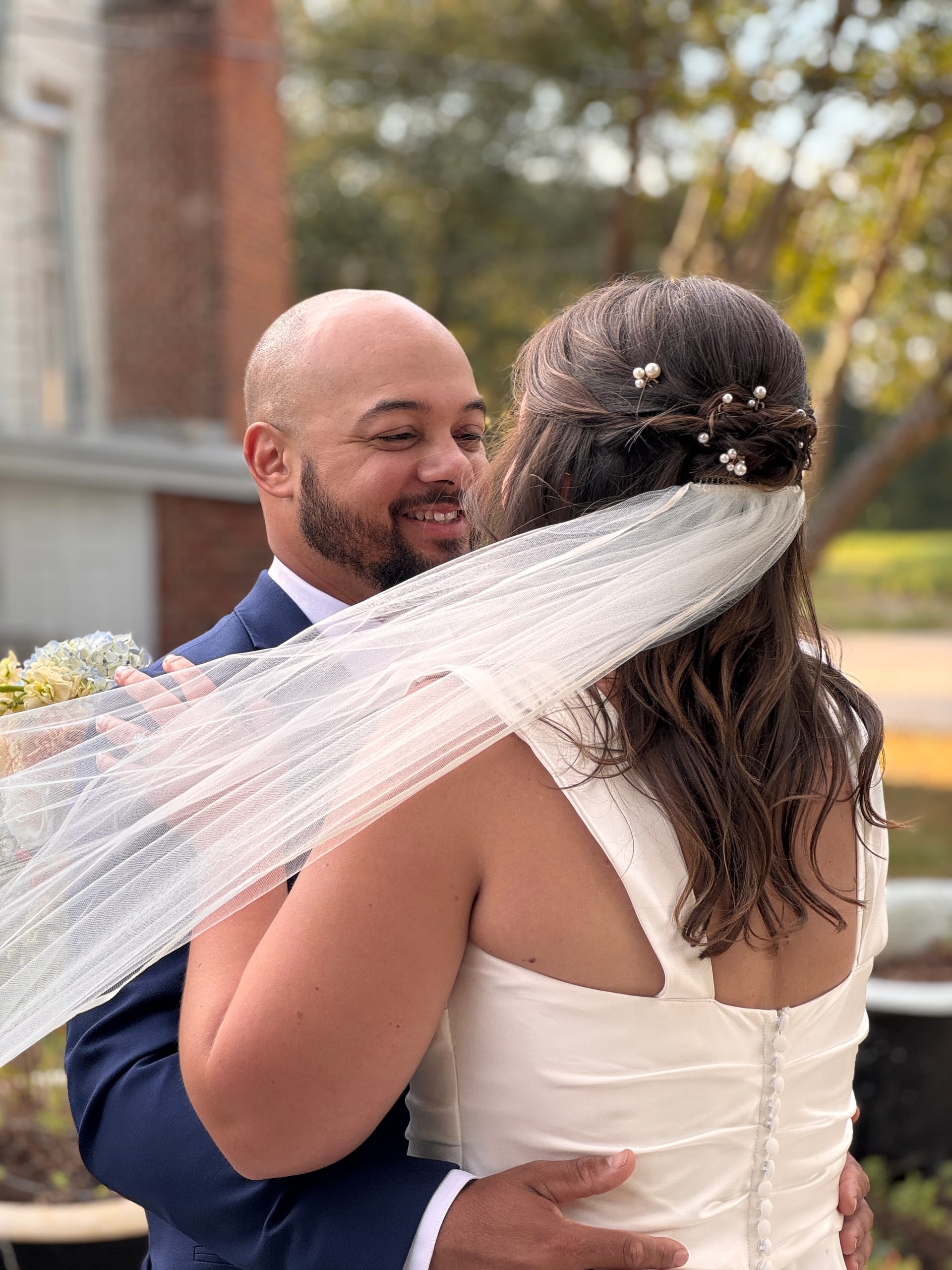 Smiling groom in a navy suit embraces his bride in a white gown and flowing veil adorned with pearl hair pins during a tender first-look moment on the ceremony lawn at Scarborough House.