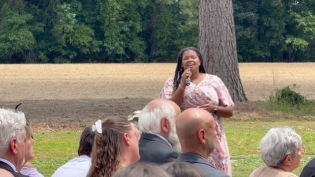 A woman in a floral dress sings into a microphone at an outdoor wedding ceremony on the historic Scarborough House estate lawn with towering trees and farmland fields in the background.