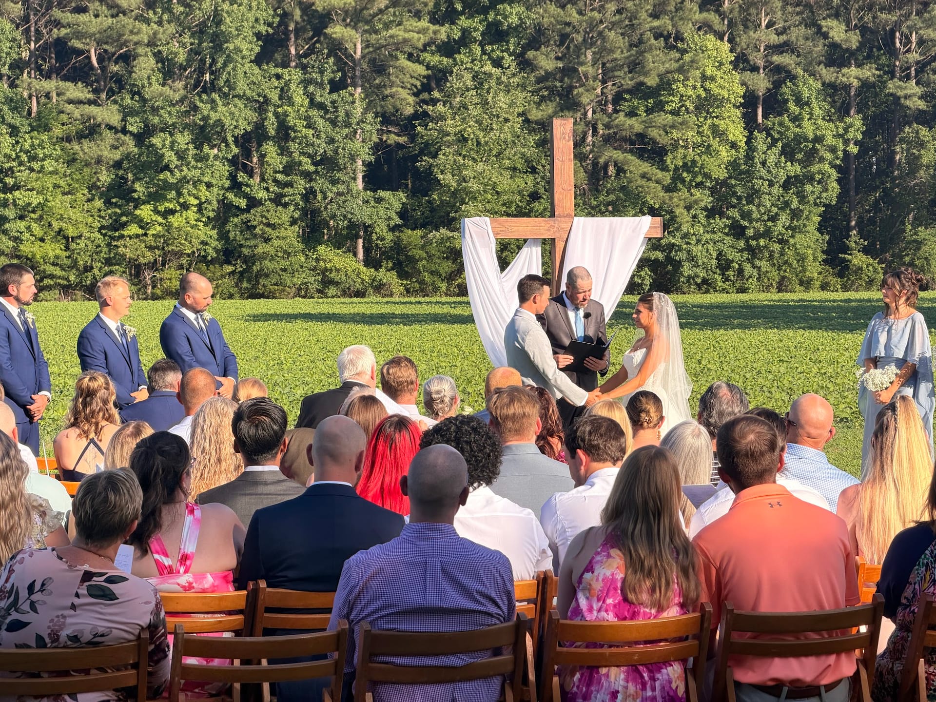 Outdoor wedding ceremony at Scarborough House with couple exchanging vows beneath a draped wooden cross altar surrounded by groomsmen and bridesmaid, with guests seated on wooden chairs overlooking green farmland fields and a forest backdrop.