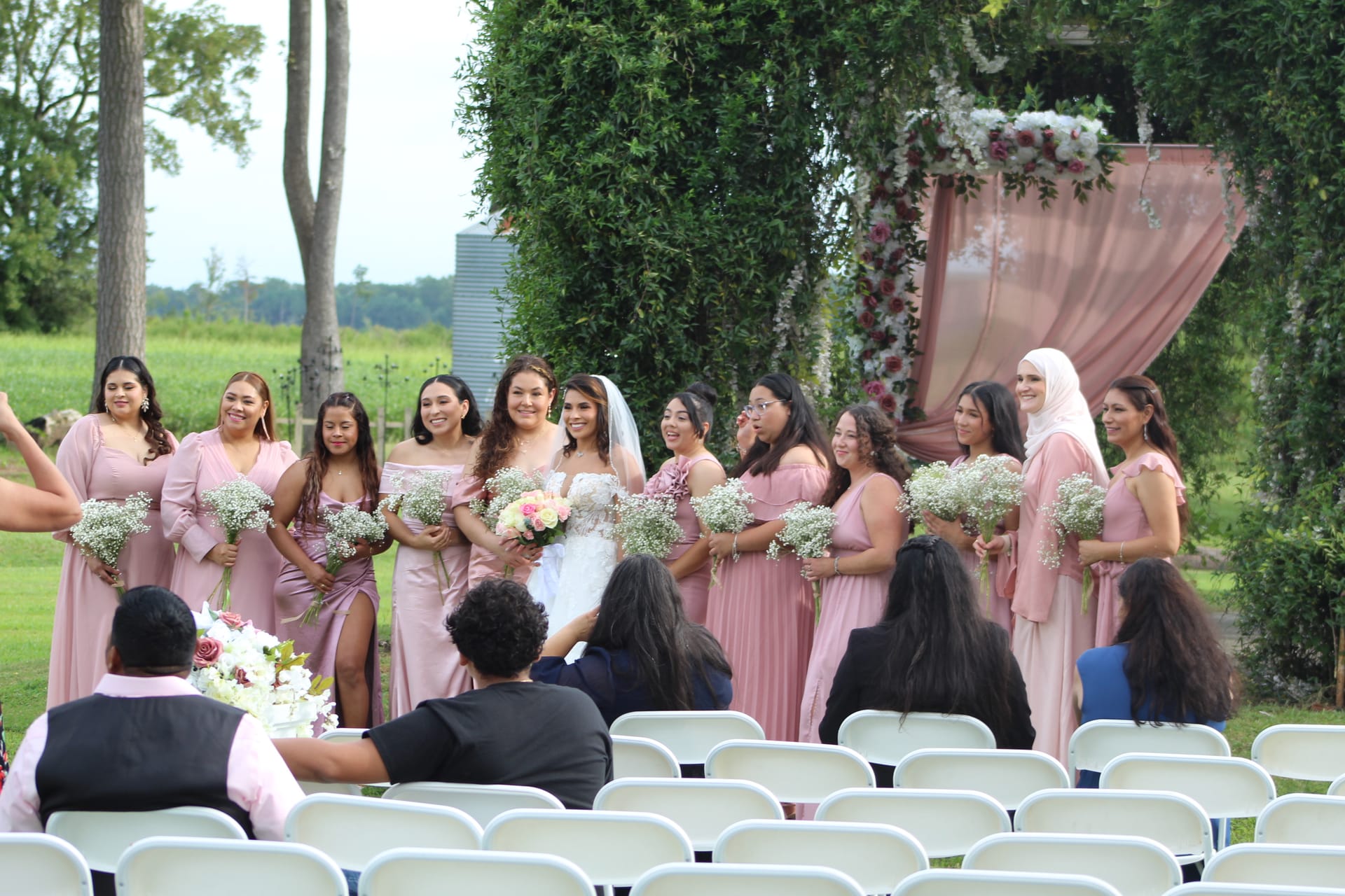 Bride in white gown and veil poses with her bridesmaids in dusty-rose dresses holding baby's breath bouquets in front of a floral and draped arch on the ceremony lawn at Scarborough House.
