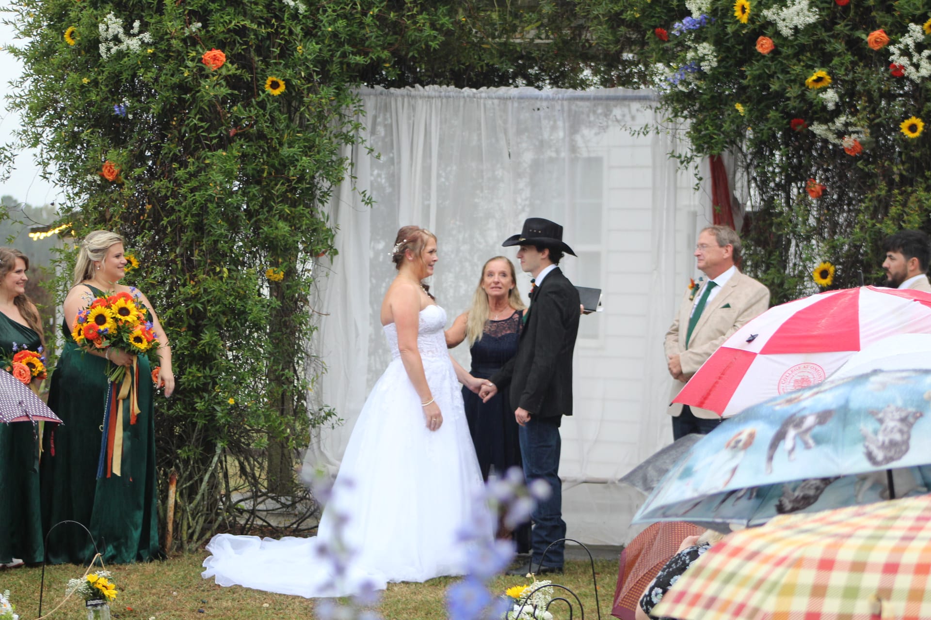 Bride and groom holding hands during outdoor vow exchange beneath a lush floral arch adorned with sunflowers and greenery at Scarborough House estate wedding venue.