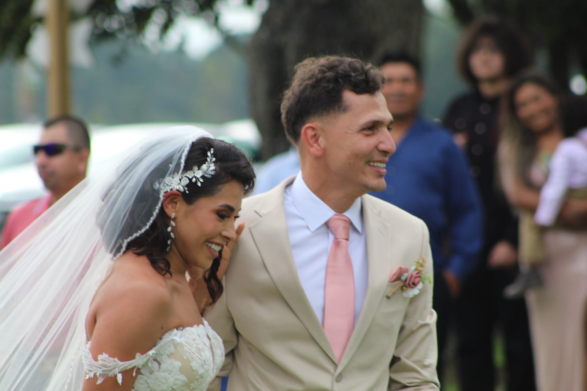 Smiling bride in lace gown and veil leans toward laughing groom in tan suit with pink tie during outdoor wedding ceremony at Scarborough House.