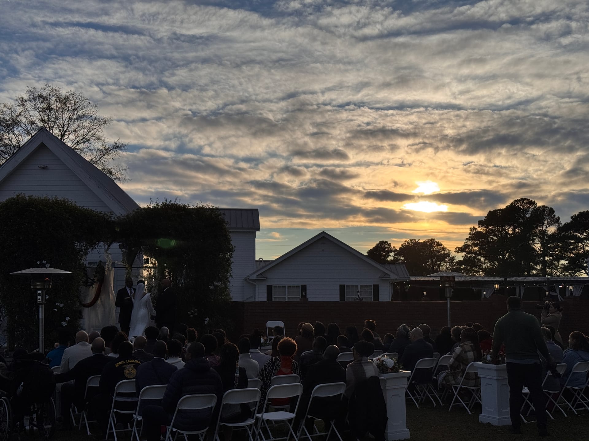 Silhouetted wedding ceremony on the outdoor lawn at Scarborough House with guests seated in rows and a breathtaking golden sunset sky overhead.
