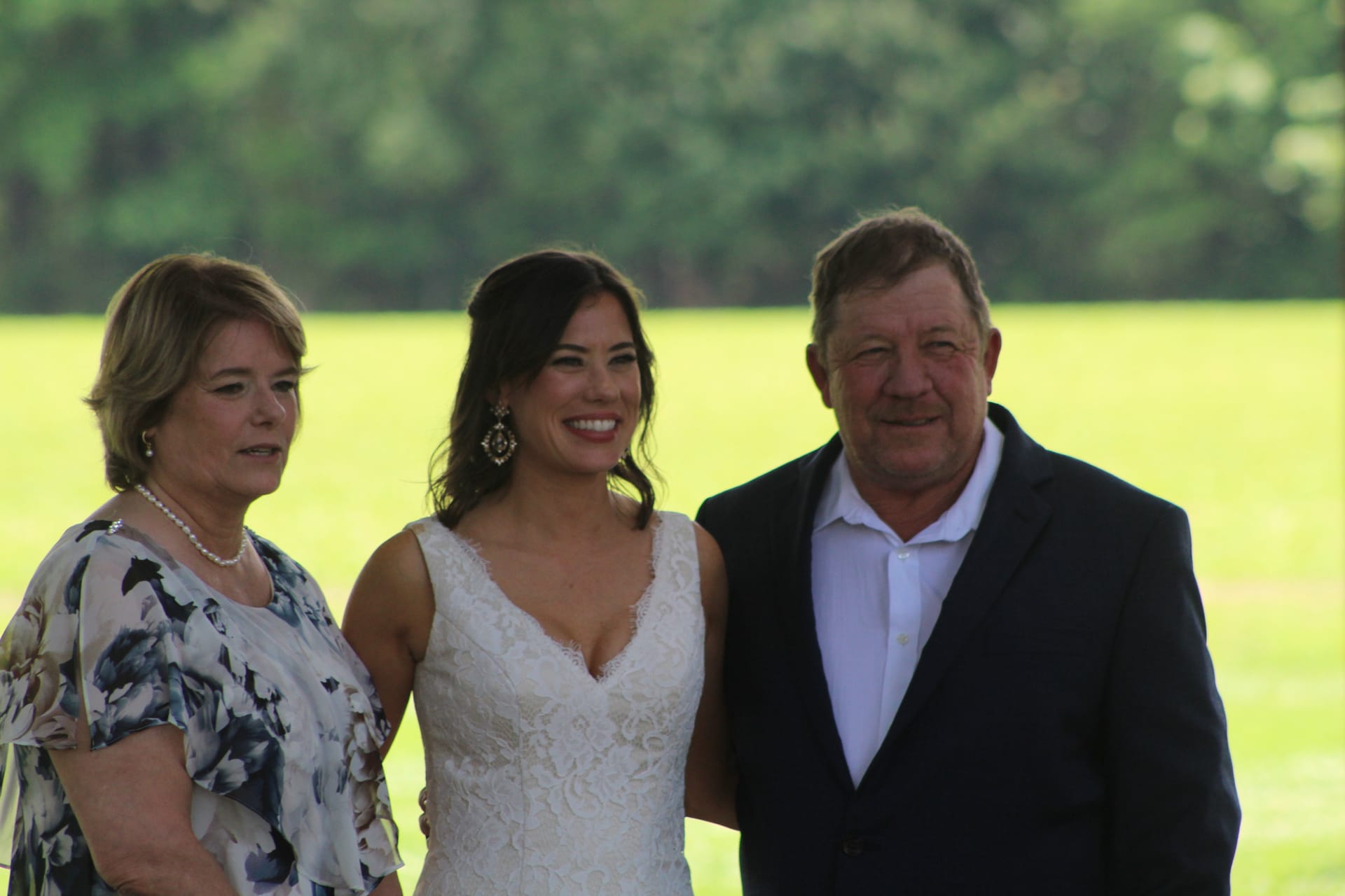 Bride in white lace wedding gown smiles with her mother and father in front of a green pastoral field at Scarborough House.