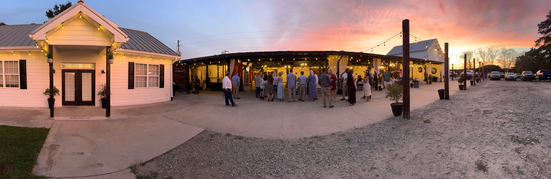 Guests mingle outside the warmly lit magnolia pavilion and white farmhouse at Scarborough House during a breathtaking pink and orange sunset cocktail hour reception.