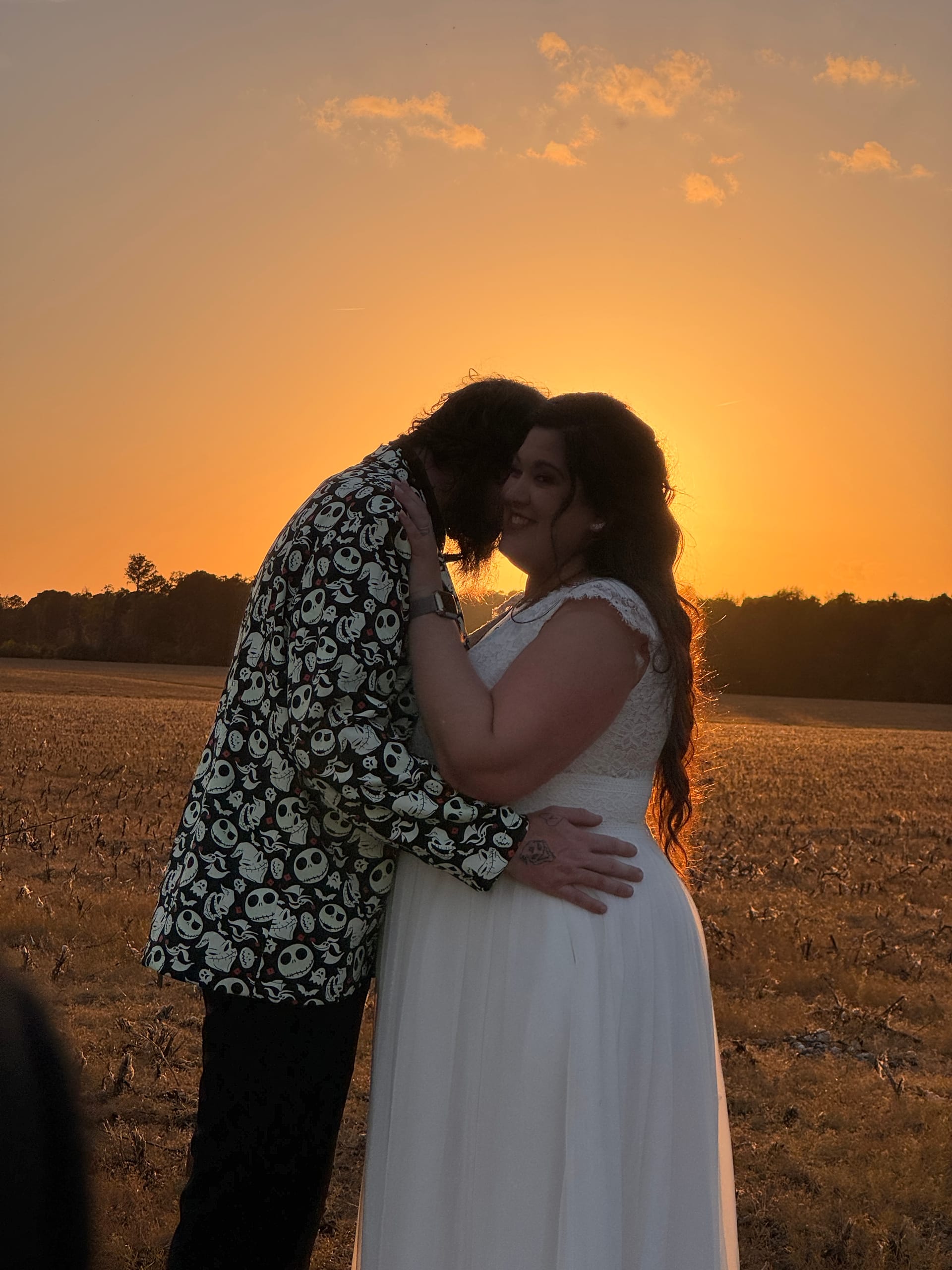 Bride in a white lace gown smiles as her groom in a patterned blazer embraces her during a vibrant orange sunset over the farmland fields at Scarborough House.