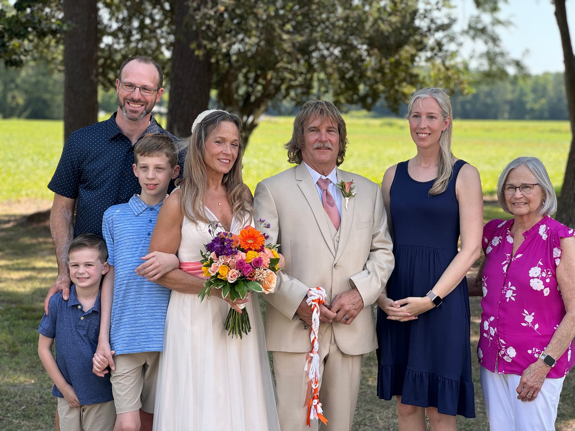 Bride and groom in a tan suit pose with family members including two young boys and an older woman beneath oak trees with open farmland fields in the background at Scarborough House.