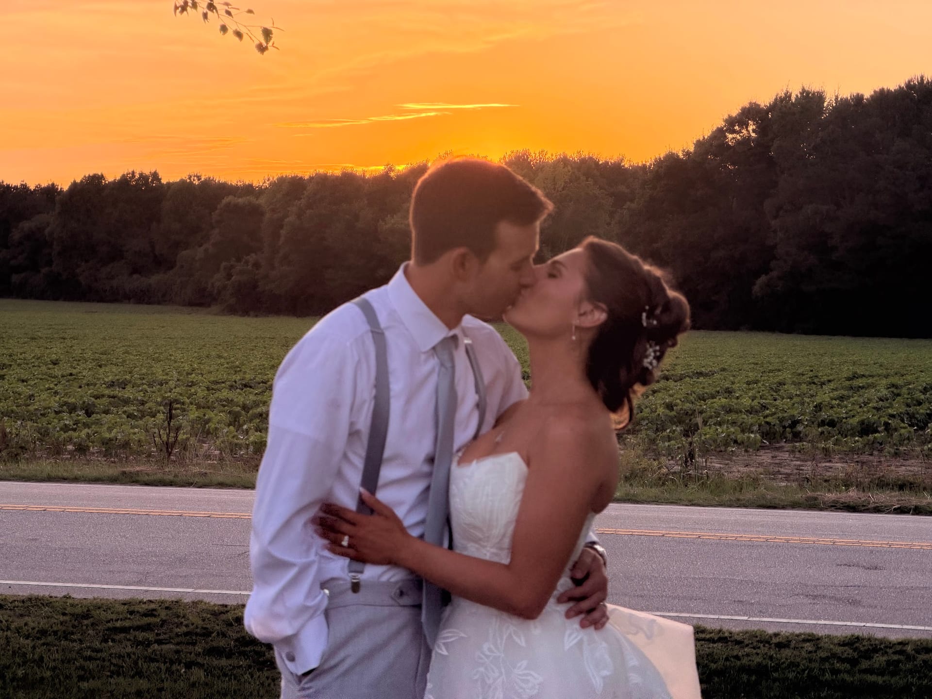 Bride and groom share a romantic sunset kiss in front of lush green farmland fields under a vivid orange sky at Scarborough House estate.