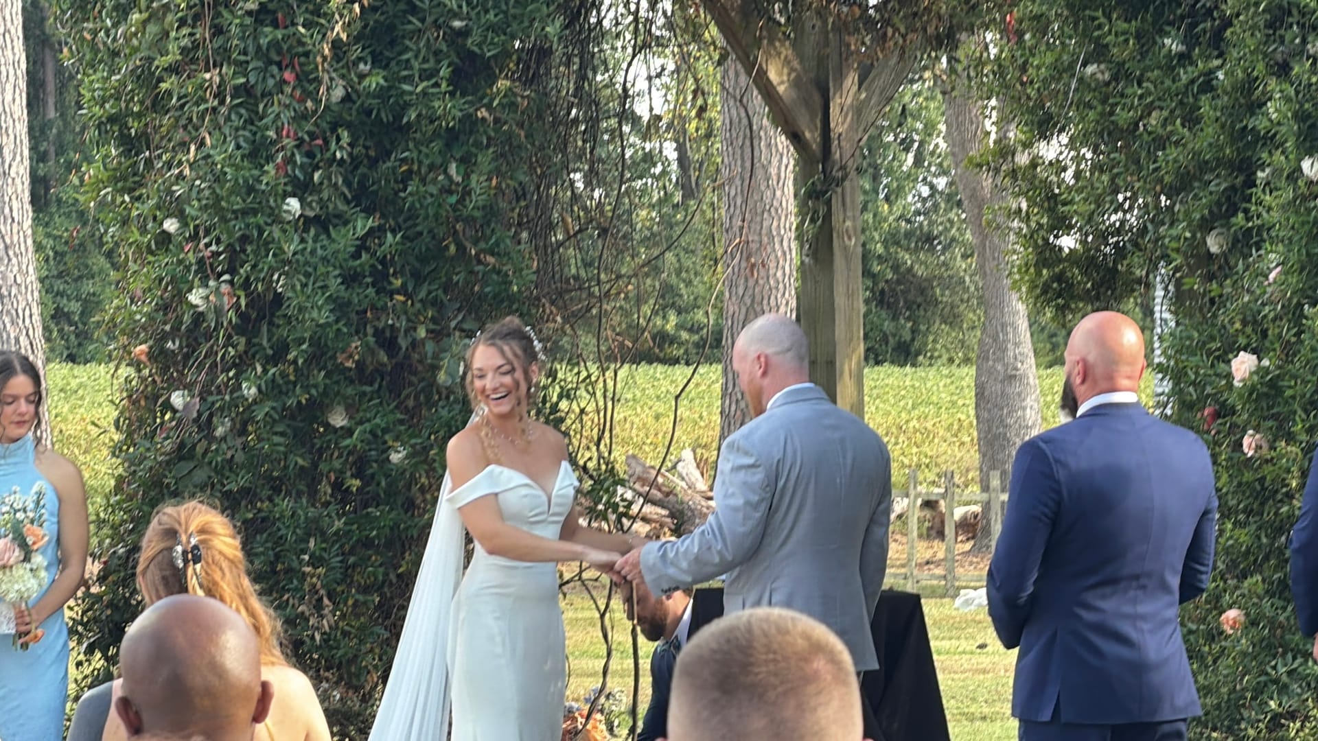 A radiant bride in an off-shoulder white gown laughs joyfully while exchanging vows with her groom on the ceremony lawn at Scarborough House, framed by tall trees and blooming greenery.