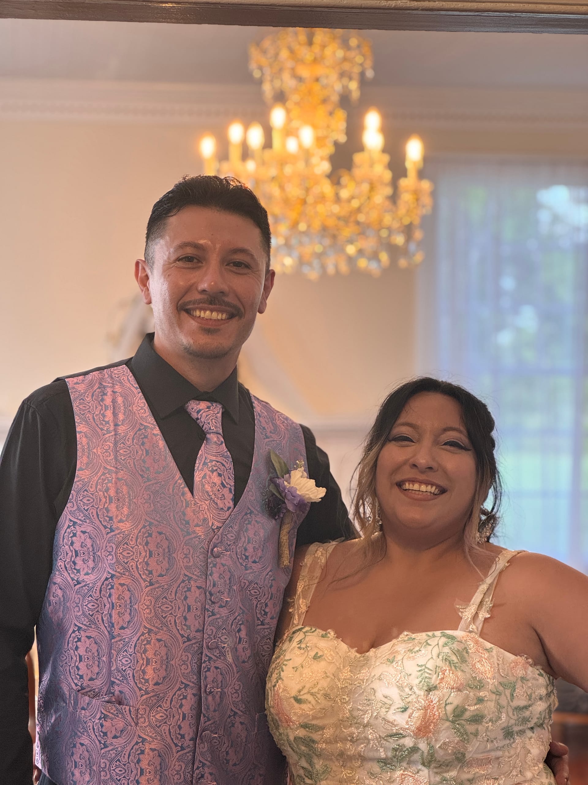 Smiling couple in wedding attire — groom in a pink paisley vest and tie and bride in a floral embroidered gown — posing beneath the golden chandelier in the grand hall of Scarborough House.