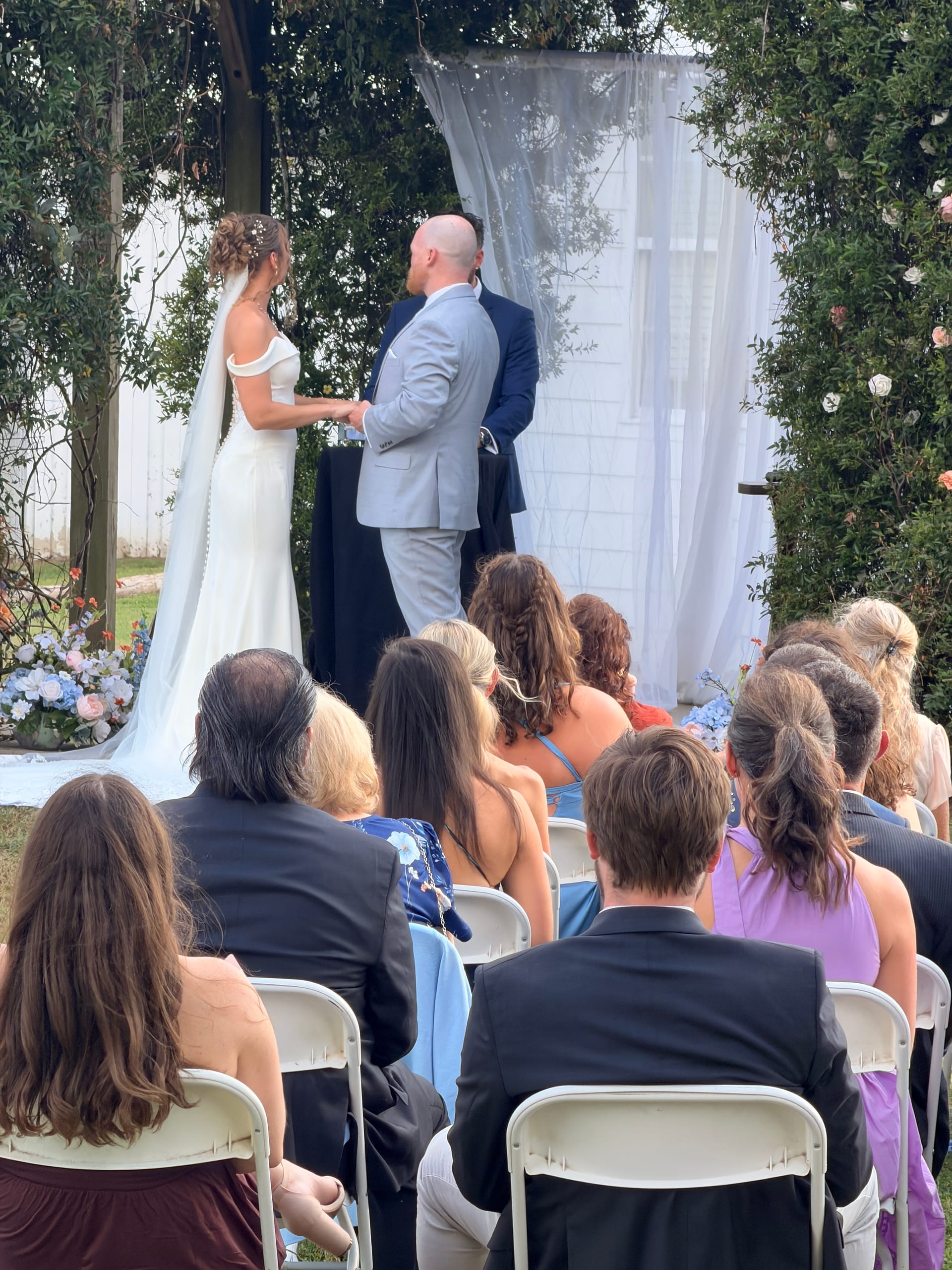 Bride and groom hold hands at a floral-adorned outdoor altar surrounded by lush greenery while seated guests watch the wedding ceremony at Scarborough House estate.