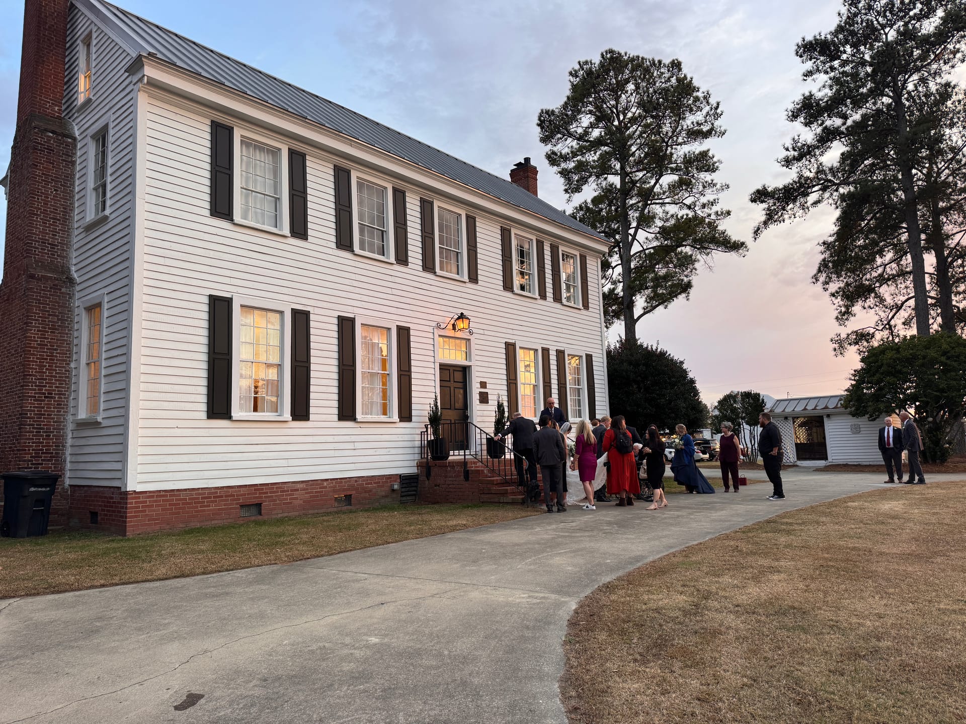 Wedding guests gather outside the illuminated 1821 Scarborough House manor at dusk, with warm light glowing through the windows and a pastel sunset sky above.
