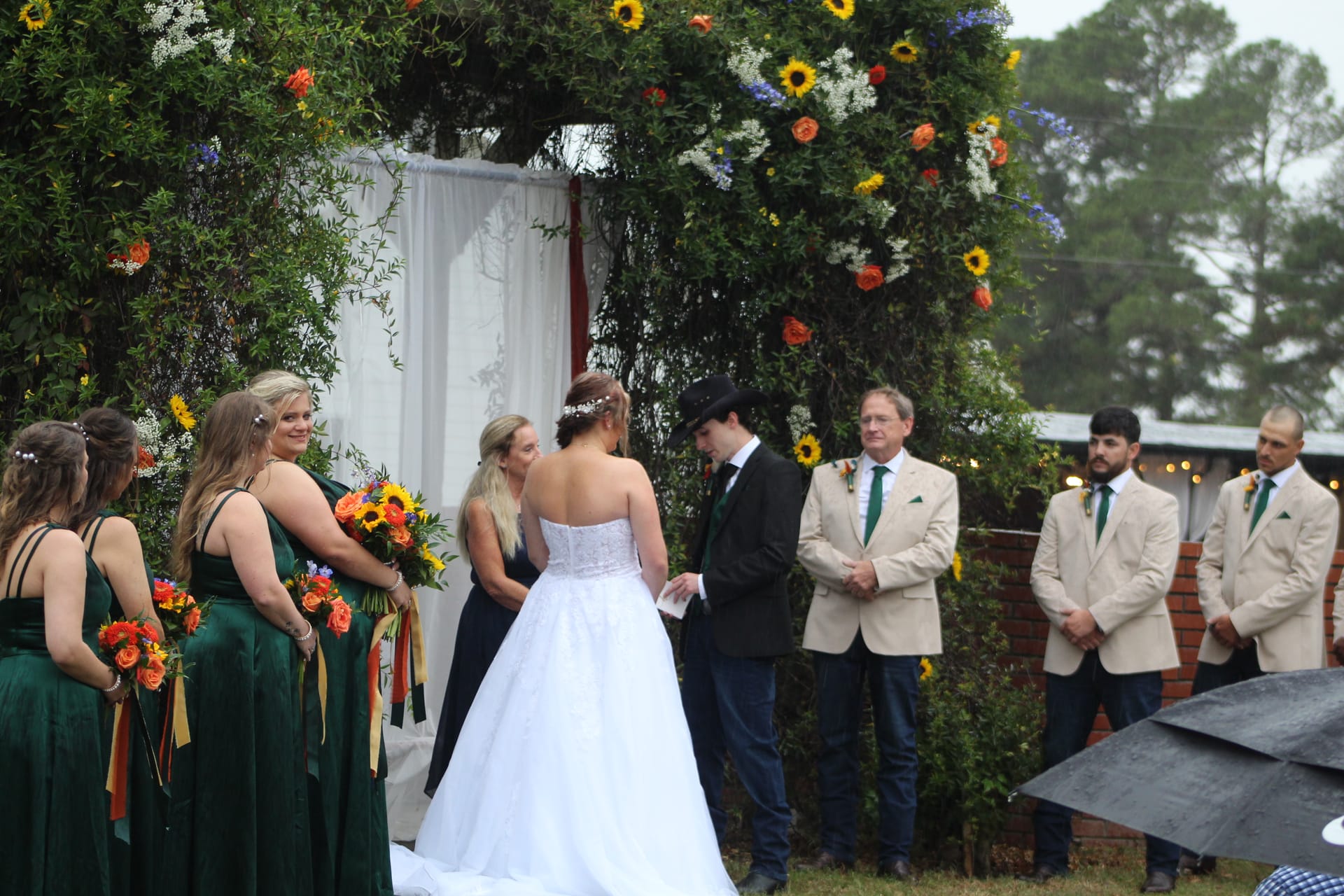 Bride and groom exchange vows beneath a lush floral arch adorned with sunflowers and orange roses as bridesmaids in emerald green dresses and groomsmen in tan jackets look on at an outdoor garden wedding ceremony.