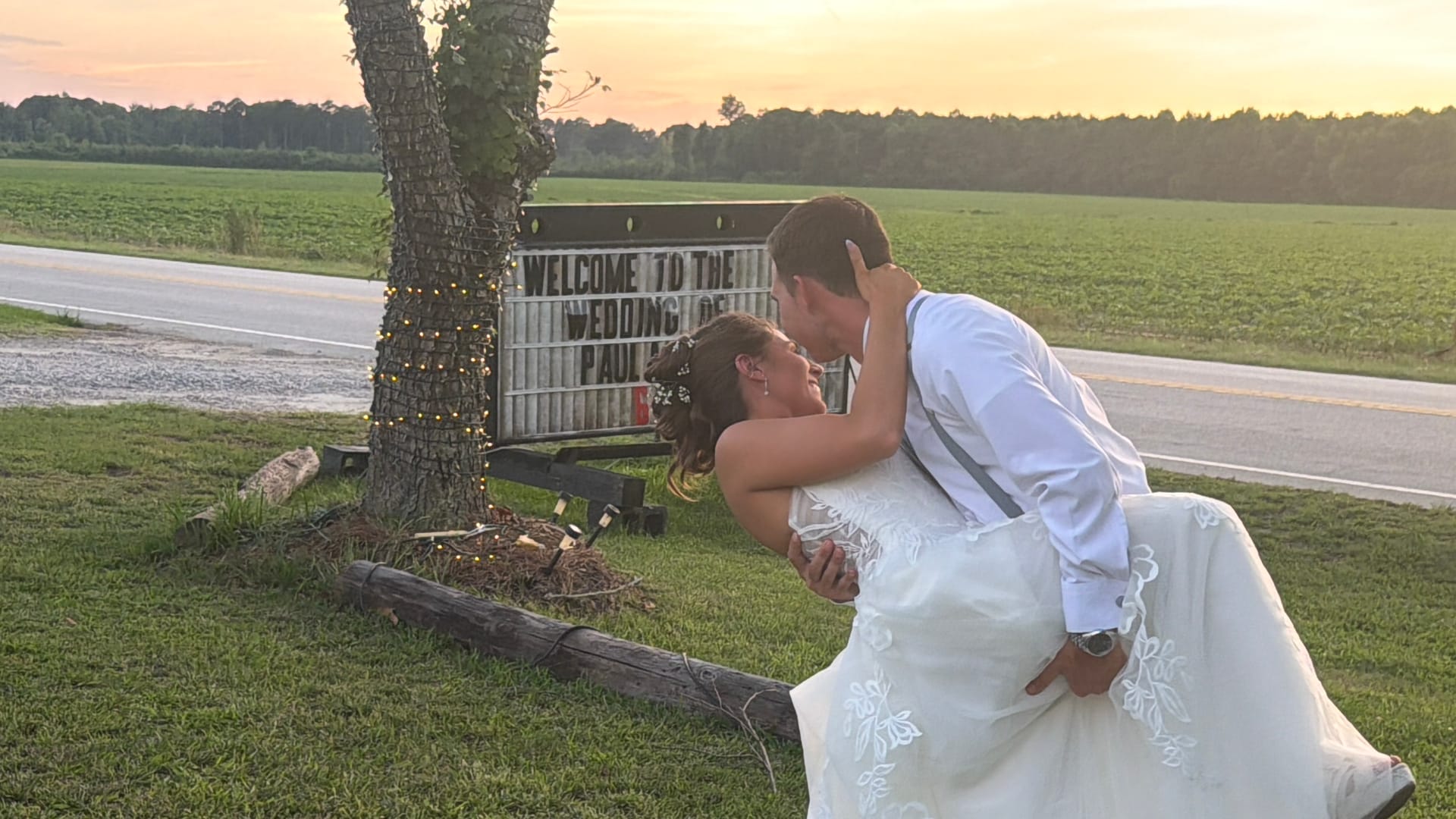 Bride in white gown being dipped and kissed by groom in suspenders beside a Welcome to the Wedding sign at sunset with open farmland fields in the background.