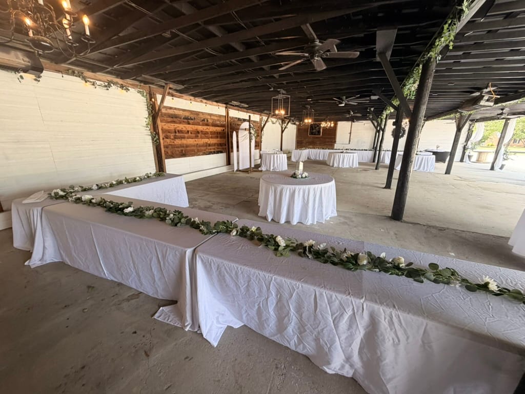 Magnolia pavilion at Scarborough House set for a wedding reception with long banquet tables draped in white linen and adorned with lush greenery and white floral garlands under a rustic wooden ceiling with chandeliers and ceiling fans.