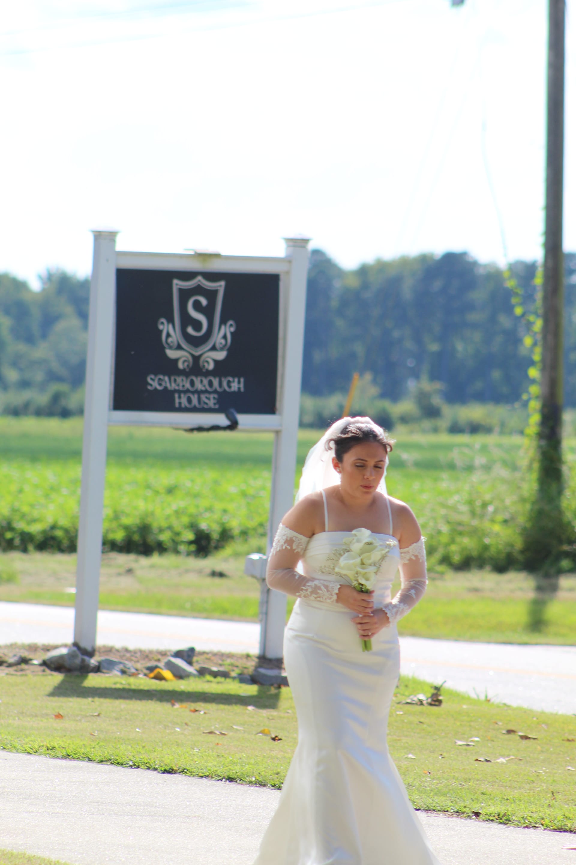Bride in a fitted off-shoulder white gown with lace sleeves holding a white calla lily bouquet walking past the Scarborough House entrance sign with green fields and trees in the background.