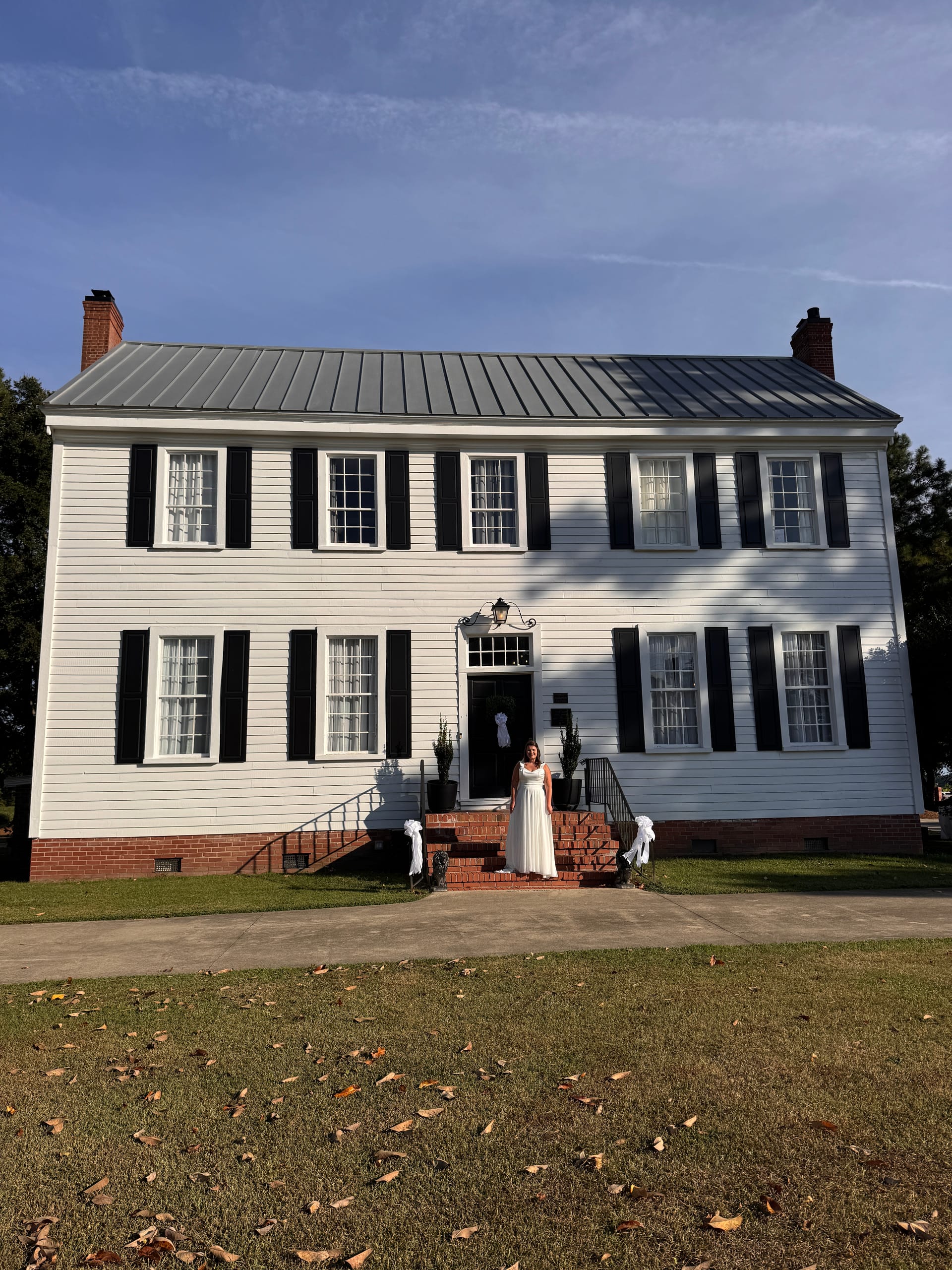 Bride in a flowing white gown standing on the brick front steps of the 1821 Scarborough House manor, a white two-story historic estate with black shutters and a metal roof under a clear blue sky.