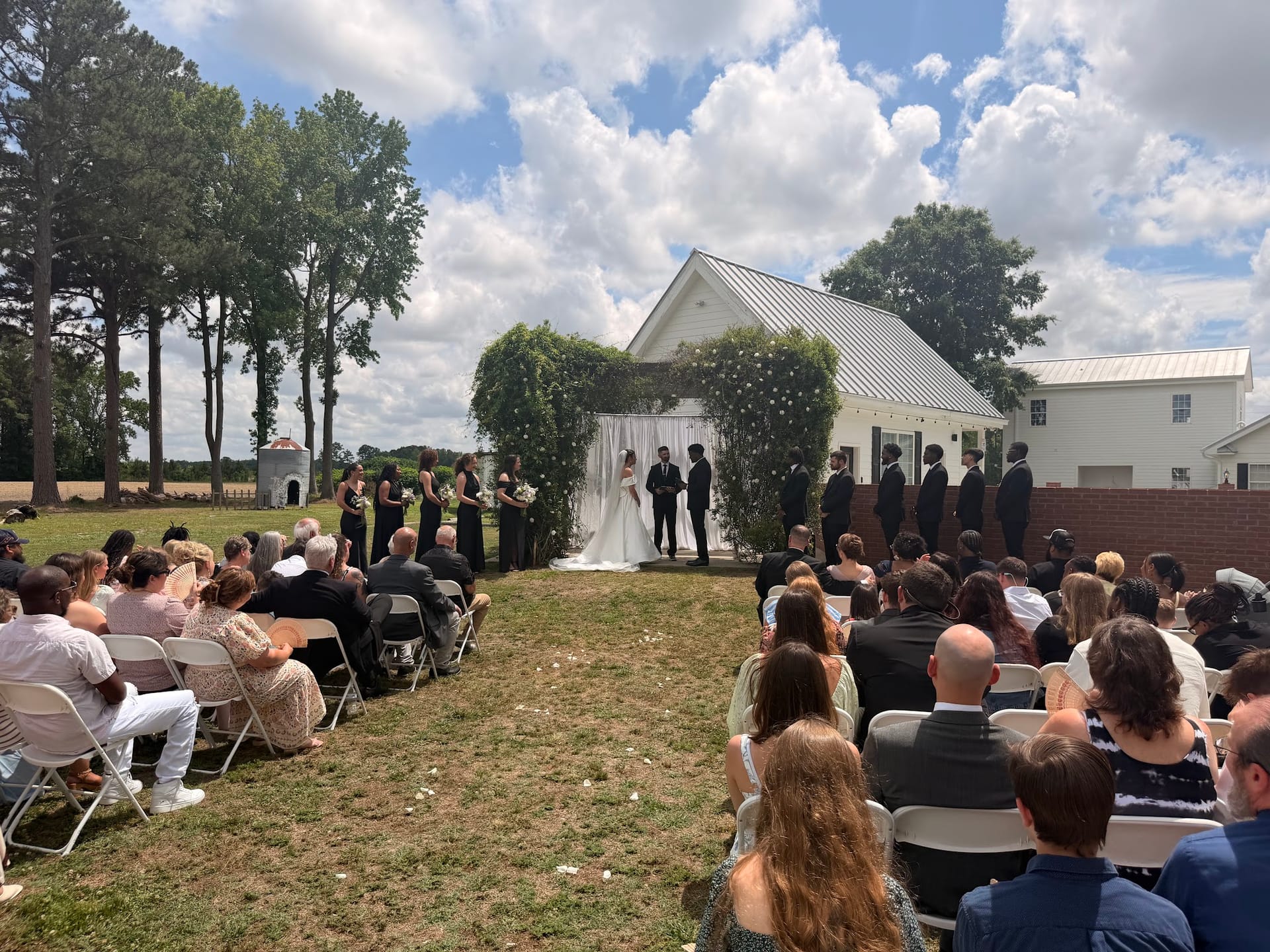 Outdoor wedding ceremony on the lawn at Scarborough House with guests seated in white folding chairs, bridesmaids in black dresses, groomsmen in dark suits, and the couple exchanging vows beneath a white floral arch in front of a vine-covered white chapel.
