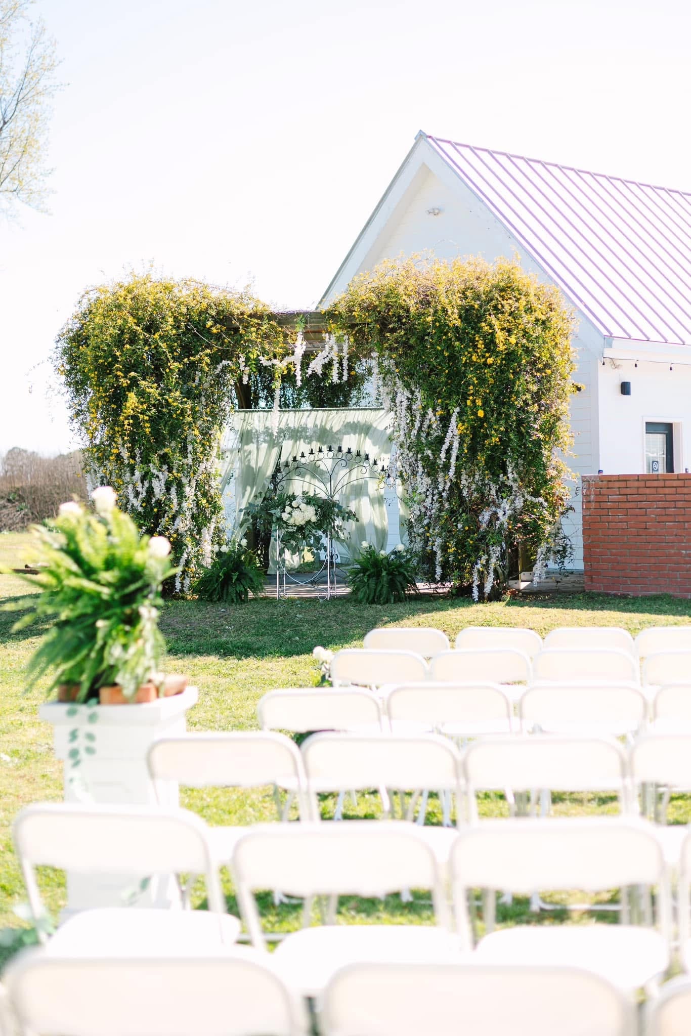 Elegant outdoor wedding ceremony setup at Scarborough House featuring rows of white folding chairs facing a lush wisteria-draped floral arch with a white manor house in the background.