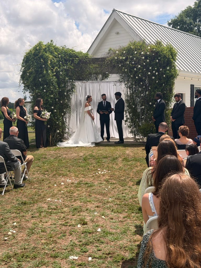 Bride and groom exchange vows beneath a lush greenery arch with white draping on the ceremony lawn at Scarborough House, surrounded by bridesmaids in black and seated guests on a sunny day.