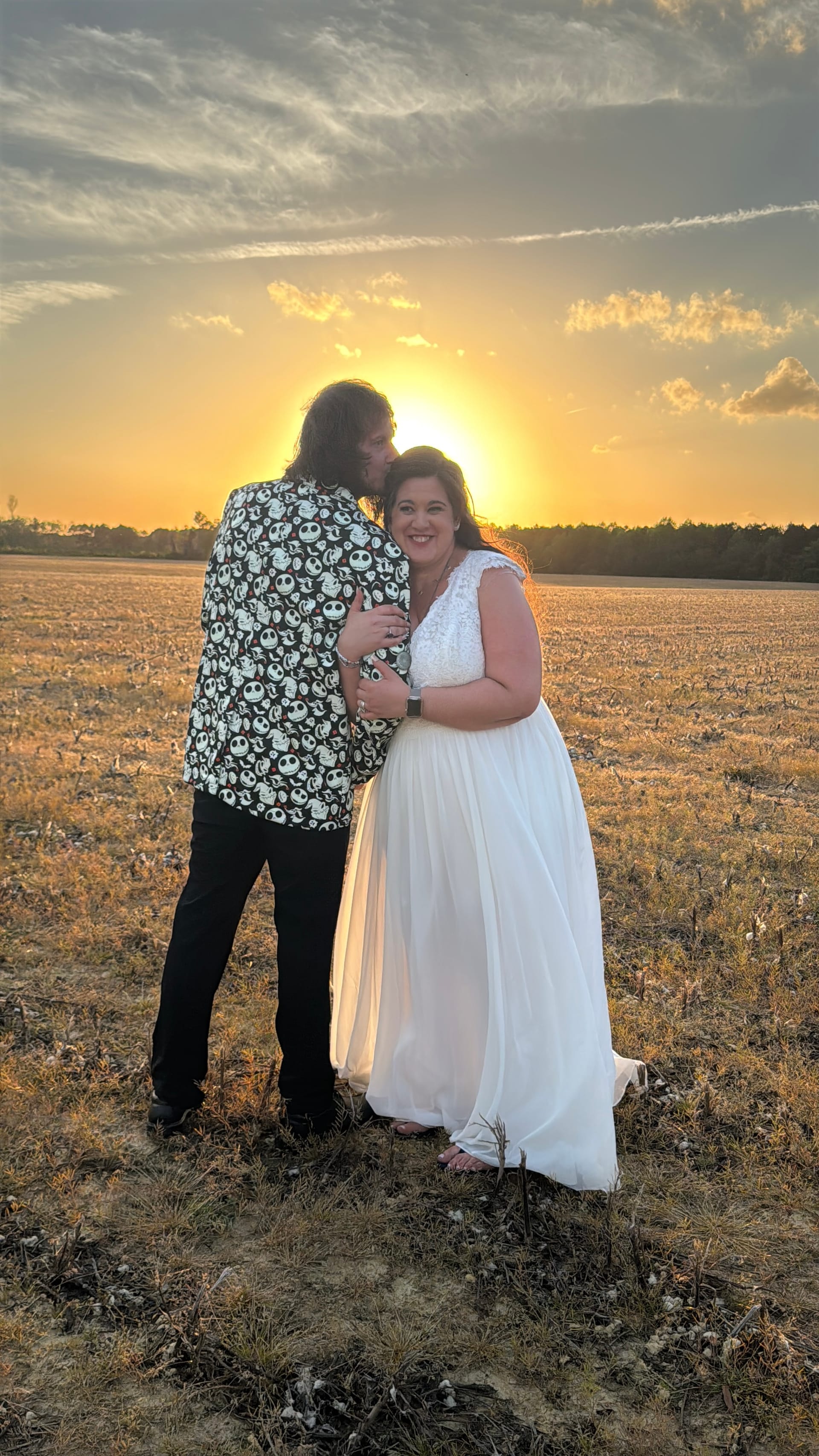 Bride in a flowing white gown smiles warmly as groom in a patterned jacket embraces her during a golden sunset over open farmland fields at Scarborough House.