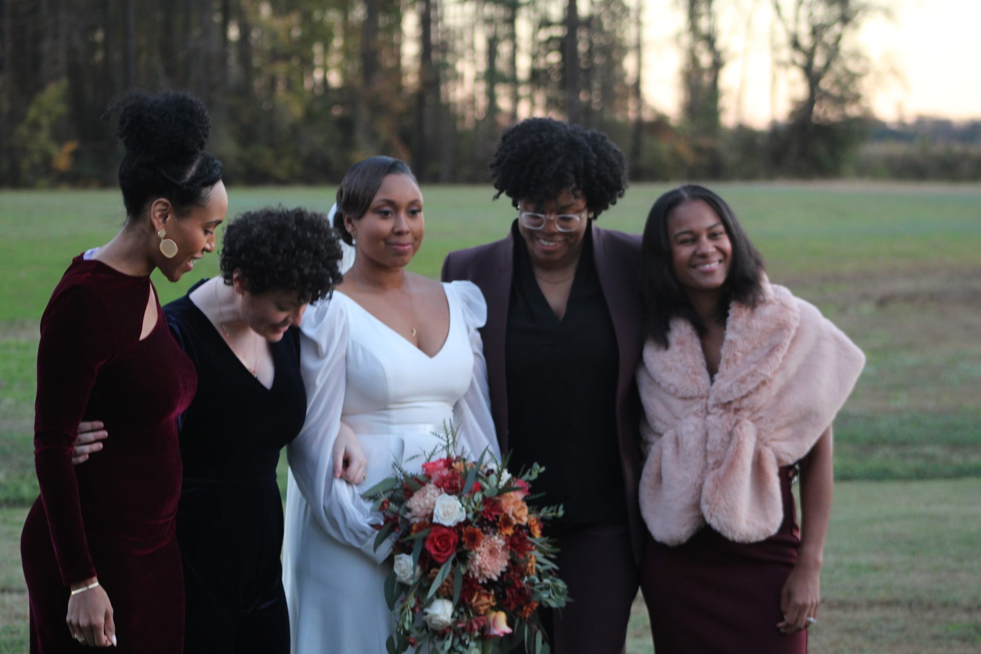 Bride in a white gown holding a colorful autumn bouquet laughs with four bridesmaids in burgundy and mauve attire on an open field at sunset.