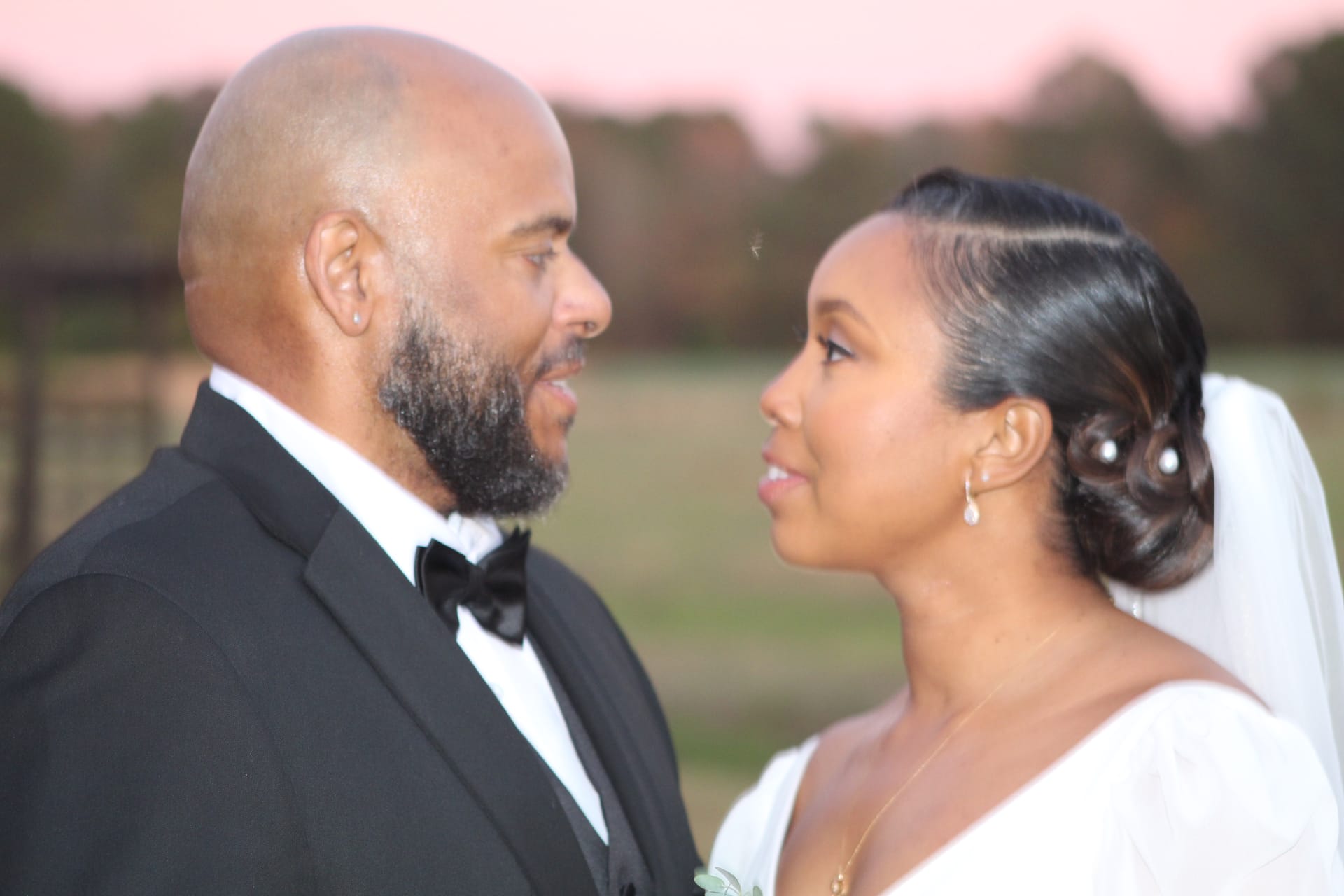 Bride in white gown and veil and groom in black tuxedo share an intimate gaze during golden hour portraits at Scarborough House estate with soft pink sunset sky and farmland fields in the background.