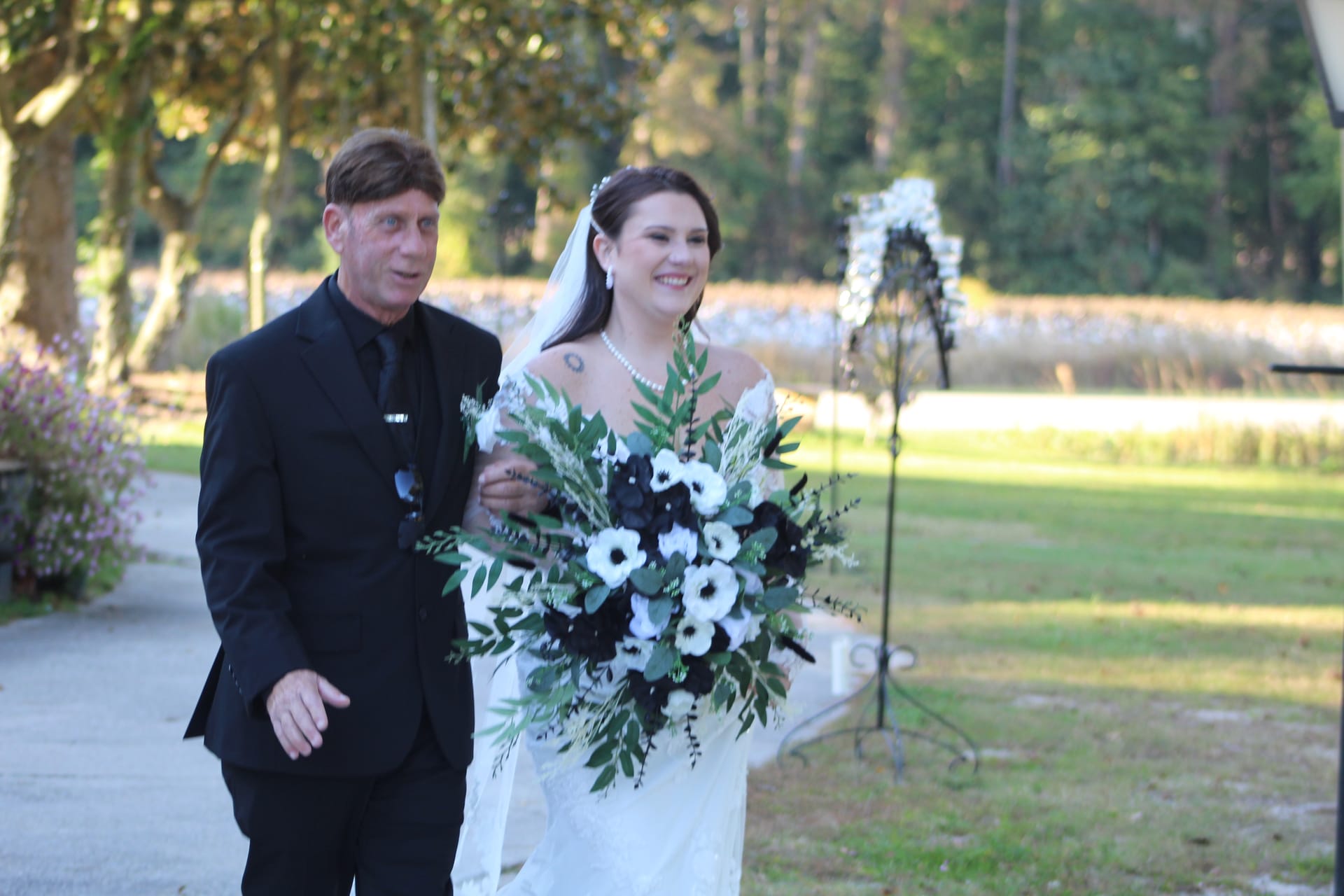 Smiling bride in a white gown carrying a dramatic black and white floral bouquet walks arm-in-arm with her escort down the outdoor aisle at Scarborough House toward a floral arch surrounded by lush trees and farmland.