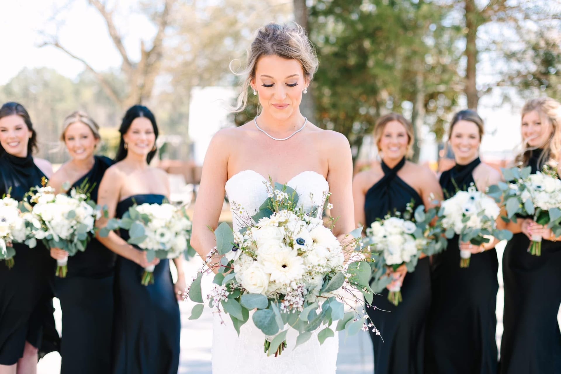 Bride in a strapless white gown holds a lush eucalyptus and white floral bouquet while her six bridesmaids in black gowns stand behind her at Scarborough House wedding venue.