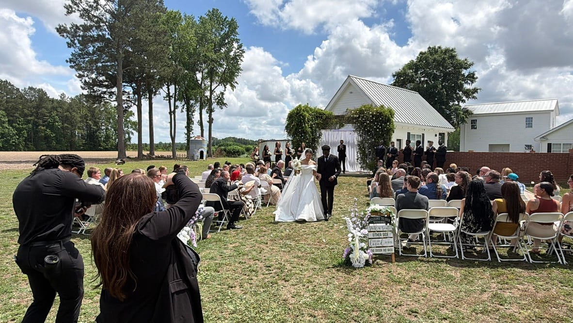 Bride and groom walk down the outdoor aisle lined with seated guests toward a floral arch on the ceremony lawn at Scarborough House historic estate on a sunny day.