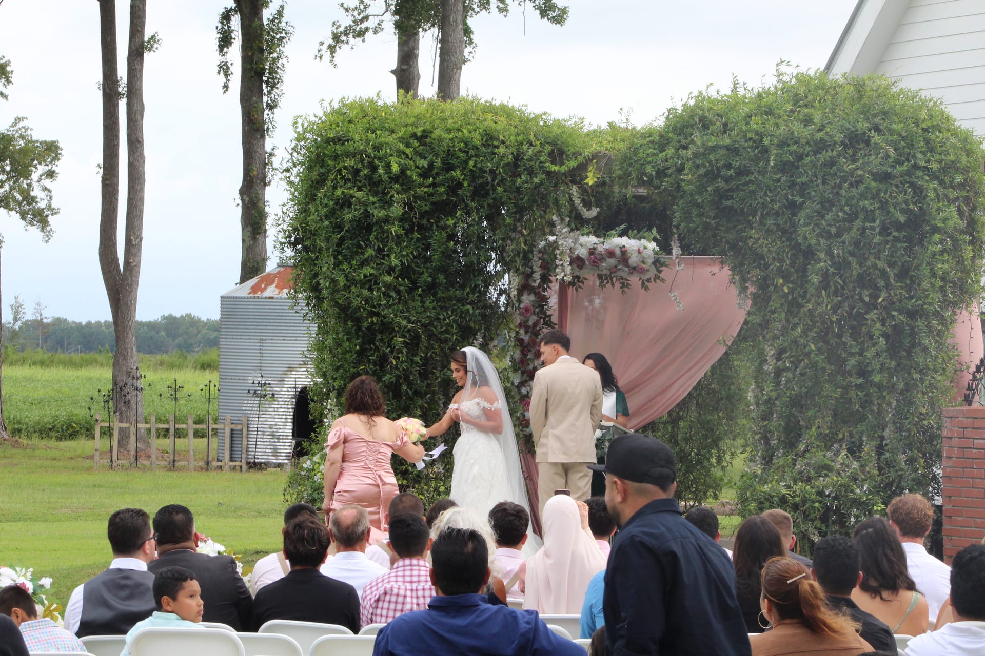 Bride and groom exchange vows beneath a lush green ivy arch adorned with blush floral arrangements and pink draping on the ceremony lawn at Scarborough House, surrounded by seated wedding guests.