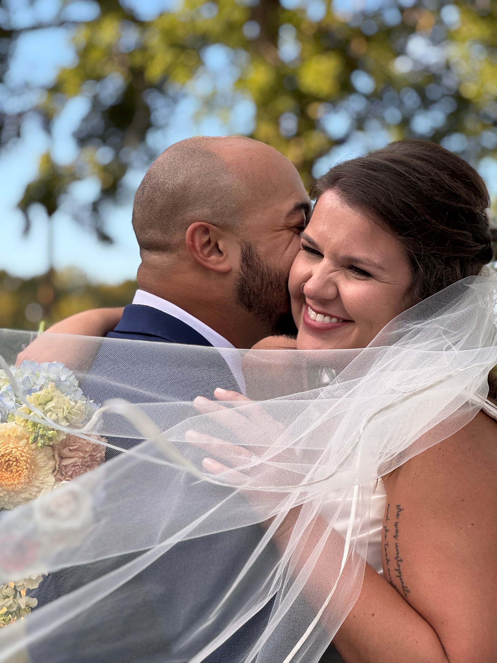 Bride laughing joyfully as groom kisses her cheek while she holds a pastel bouquet beneath oak trees at Scarborough House.