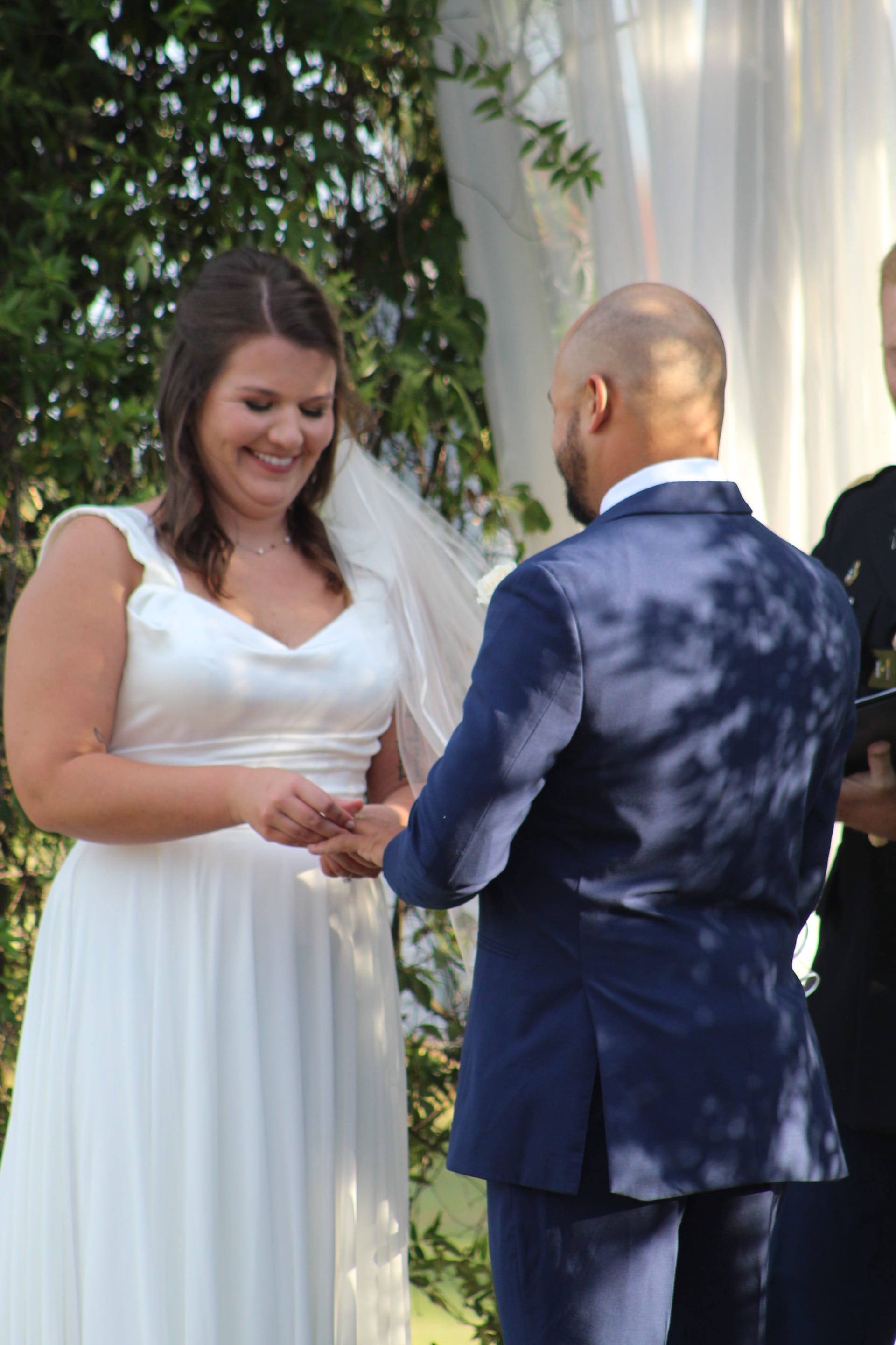 Bride in a flowing white gown smiles joyfully as groom places a ring on her finger during an outdoor wedding ceremony at a historic estate venue.