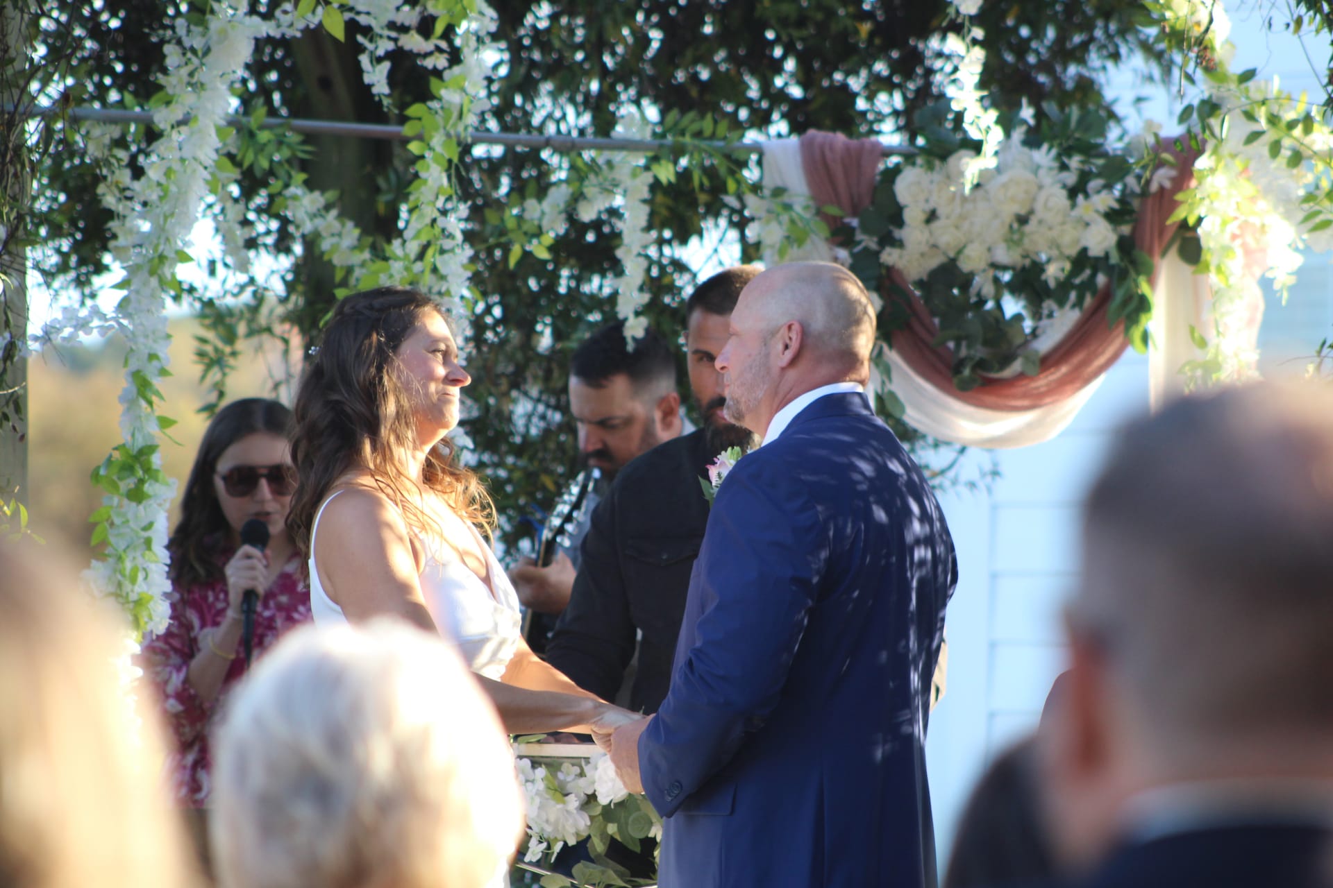 Bride and groom holding hands and exchanging vows beneath a white floral and greenery arch at an outdoor wedding ceremony at Scarborough House estate.