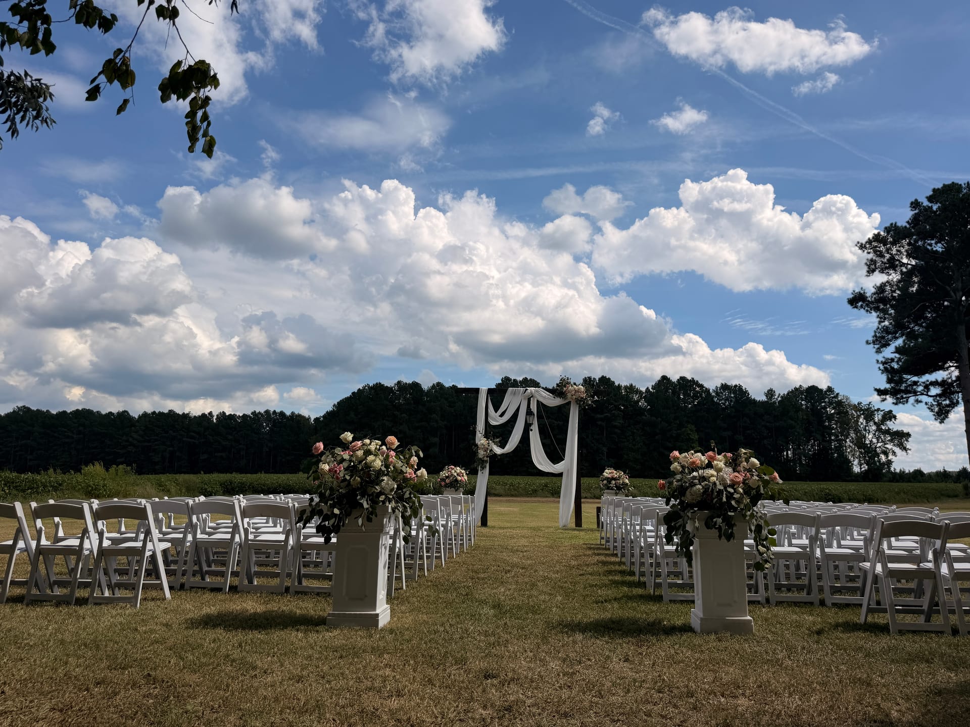 Outdoor wedding ceremony setup at Scarborough House featuring white folding chairs arranged along a grass aisle with lush floral pedestals and a draped wooden arch against a dramatic blue sky.